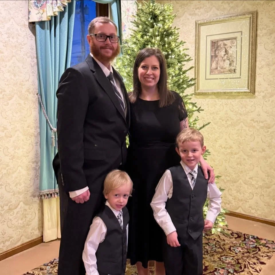 A family of four standing in front of a decorated Christmas tree in a living room. The father, mother, and two young boys are dressed formally, smiling at the camera.