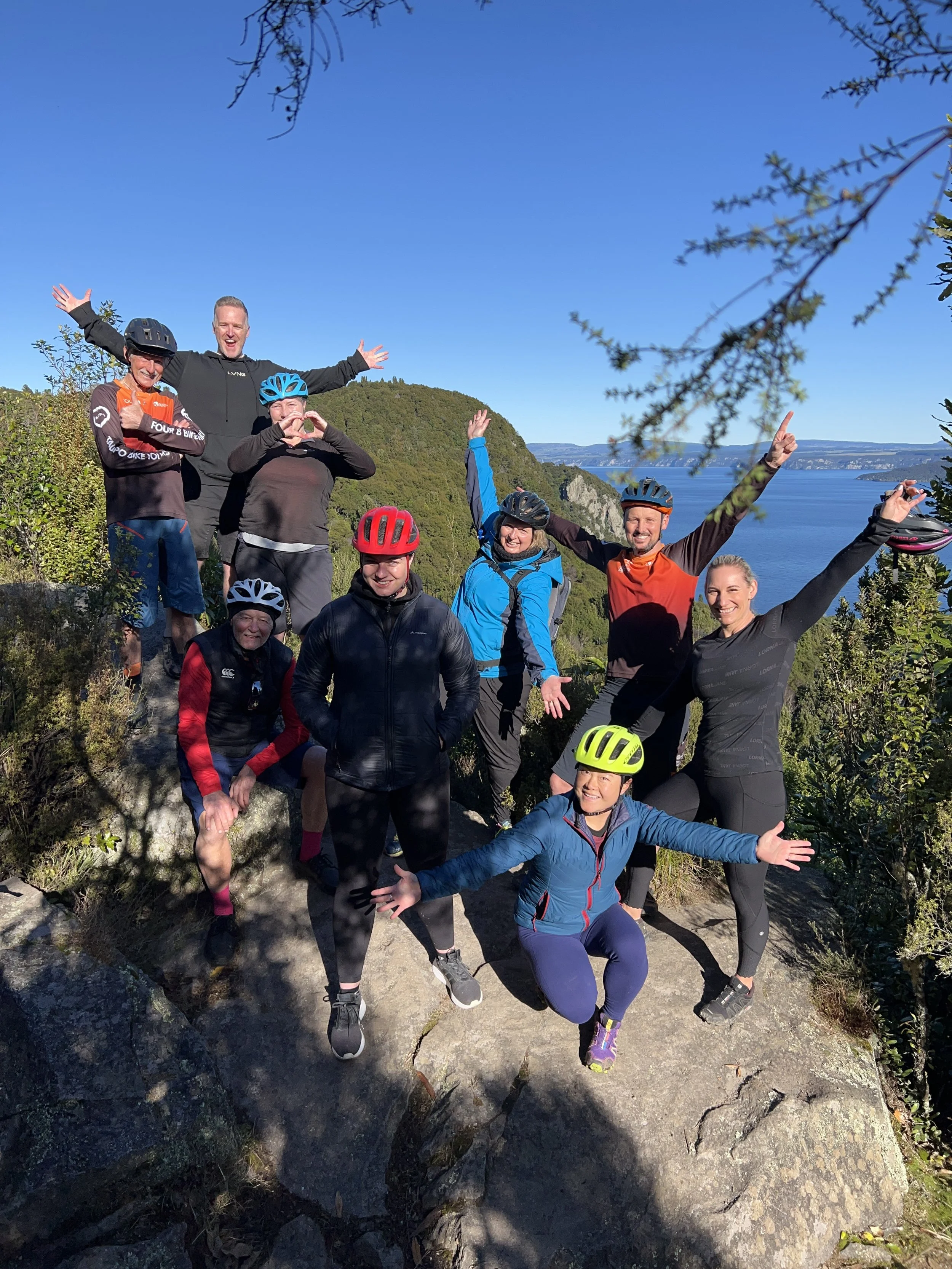 Group of nine people posing on a rocky trail with ocean and hills in the background, some wearing helmets and athletic clothing, celebrating outdoor adventure.