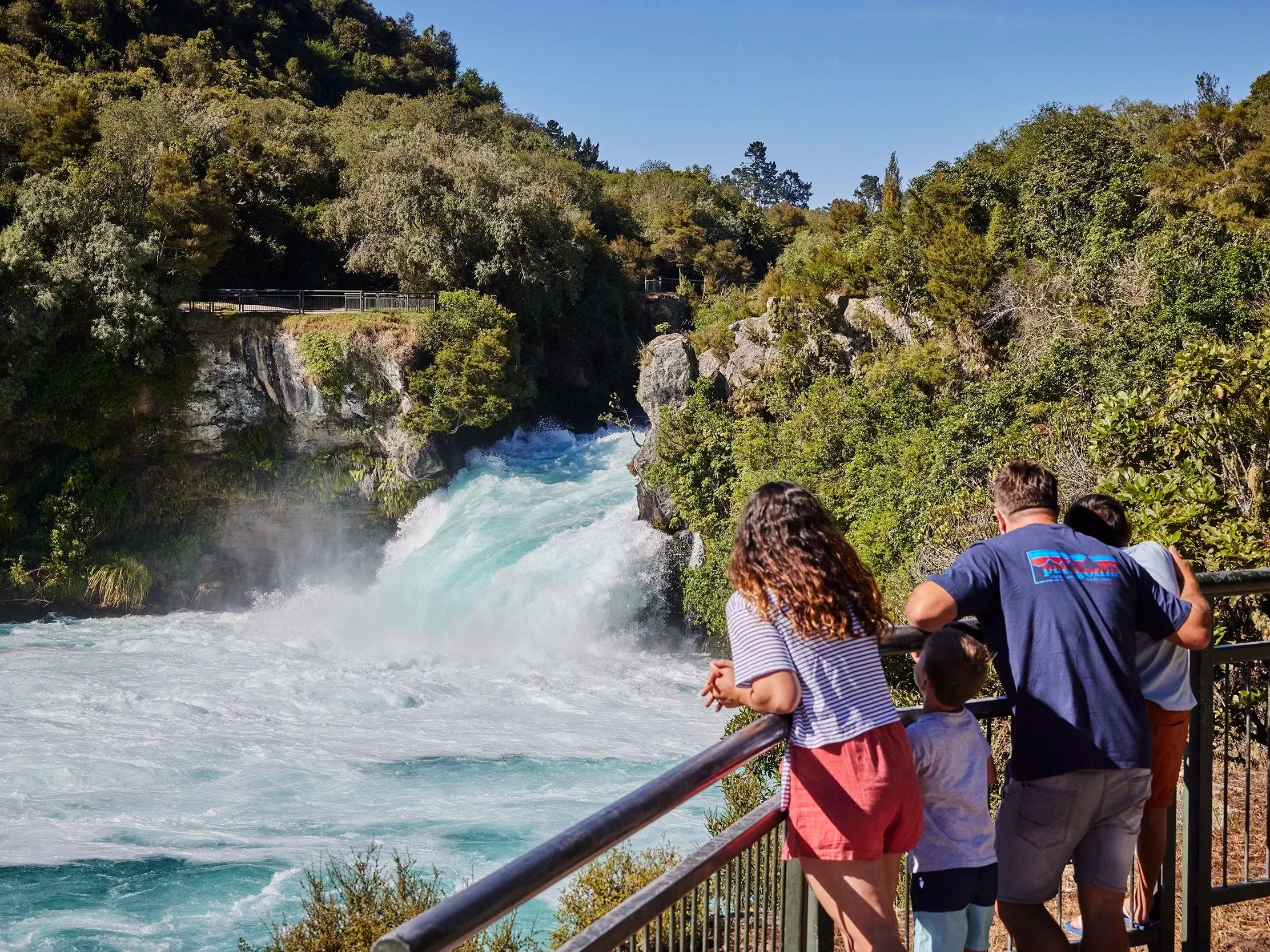 People standing on a bridge overlooking a rushing river surrounded by lush green trees and hills under a clear blue sky.