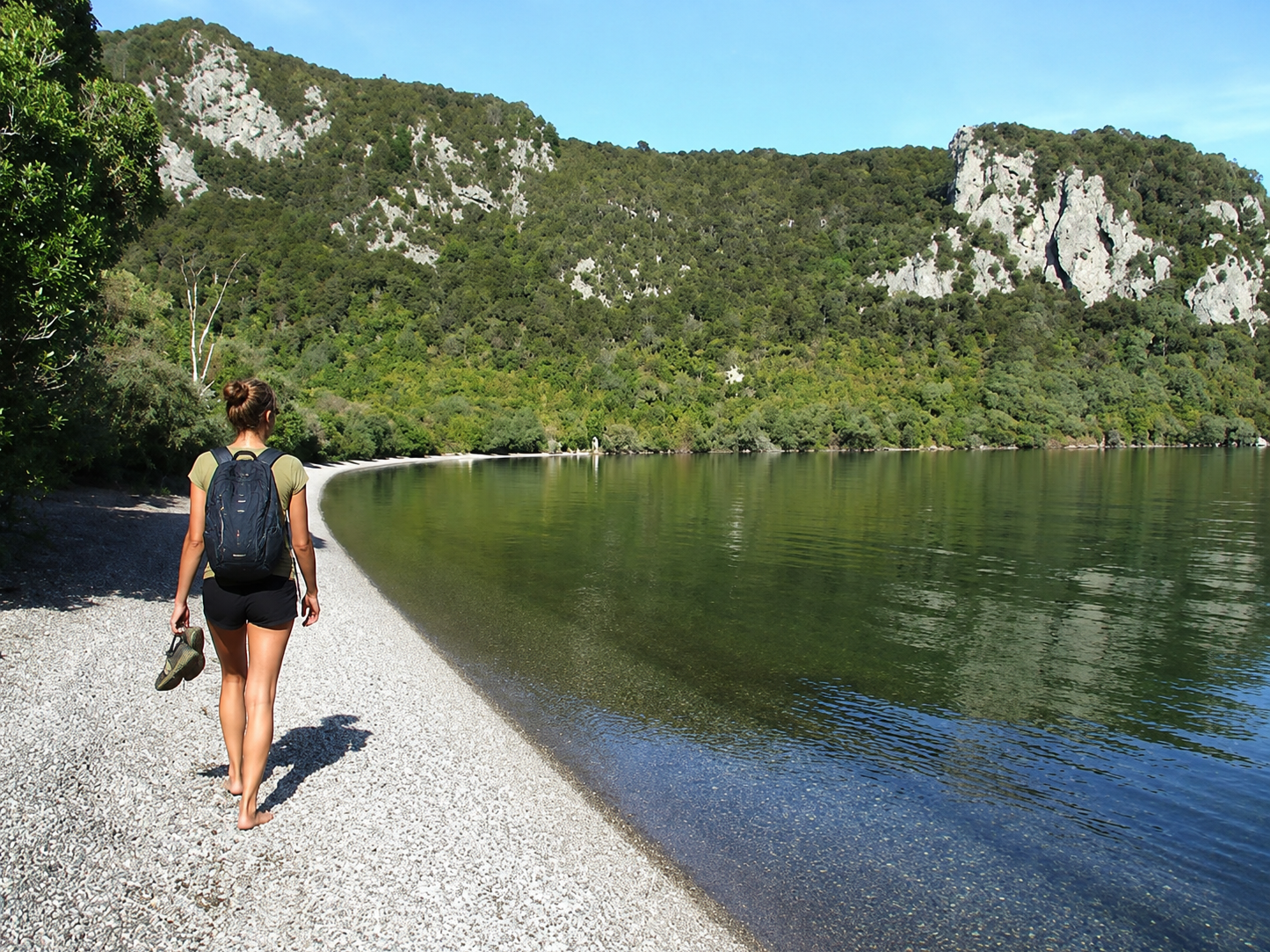 A woman with a backpack walking barefoot along a lakeside gravel path with a forested mountain in the background.