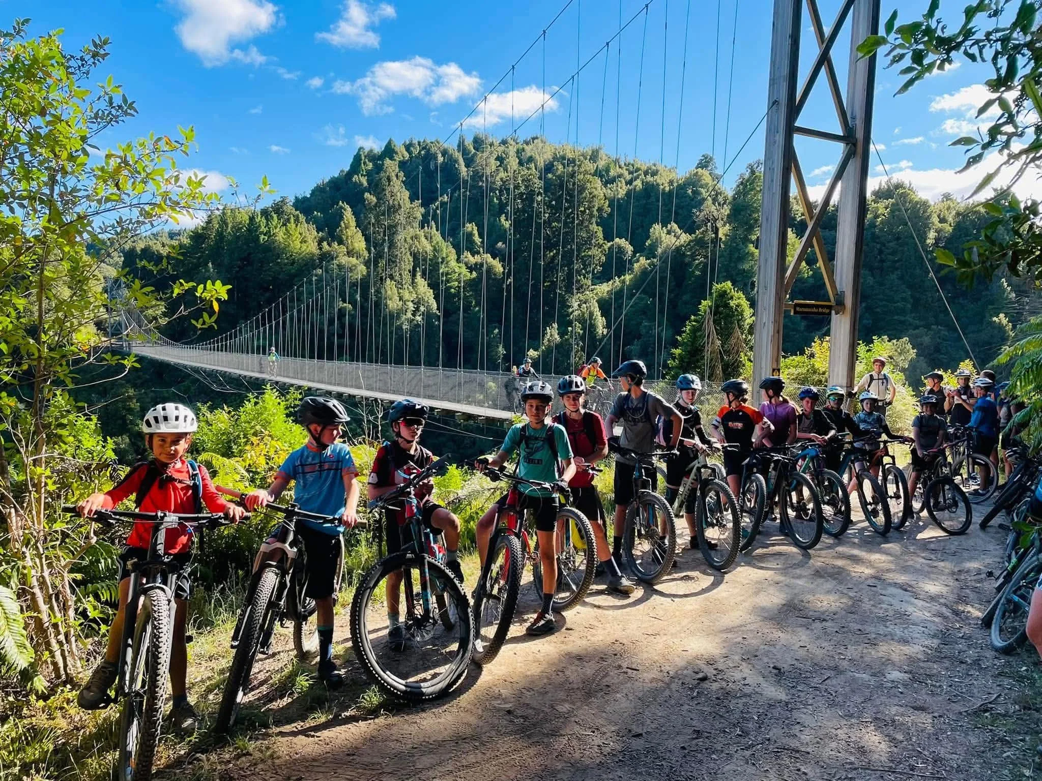 Group of children with helmets riding bicycles on a trail near a suspension bridge surrounded by trees and hills under a blue sky.