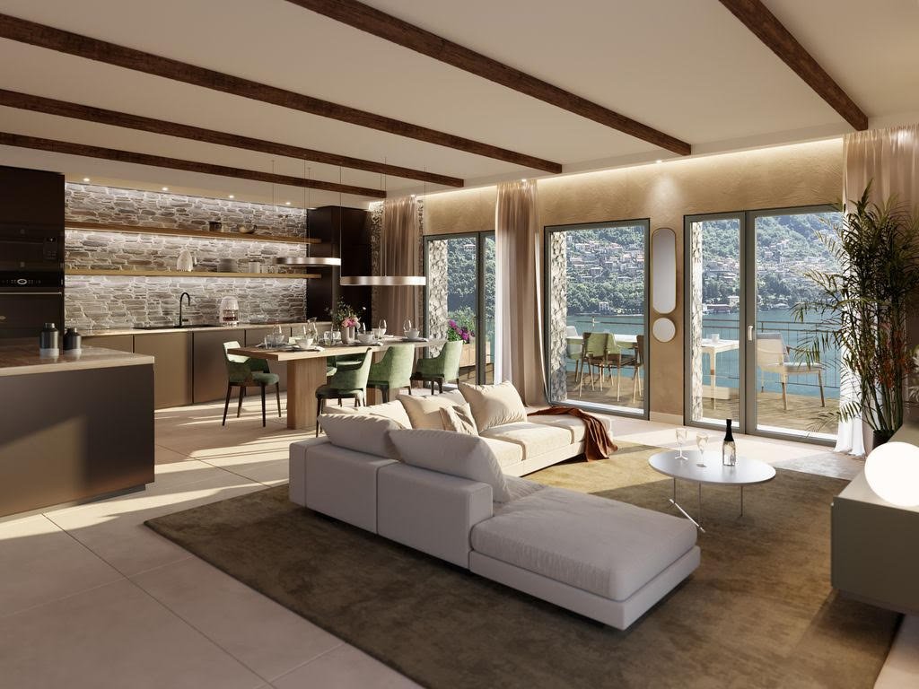 Modern living room with large windows overlooking a lake, featuring a white sectional sofa, a round coffee table, a dining area with chairs, and a balcony with outdoor furniture. The room has a neutral color palette, exposed ceiling beams, and a stone accent wall.