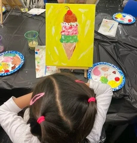 Child with pigtails painting a colorful ice cream cone on a yellow canvas, surrounded by painting supplies and plates of paint.
