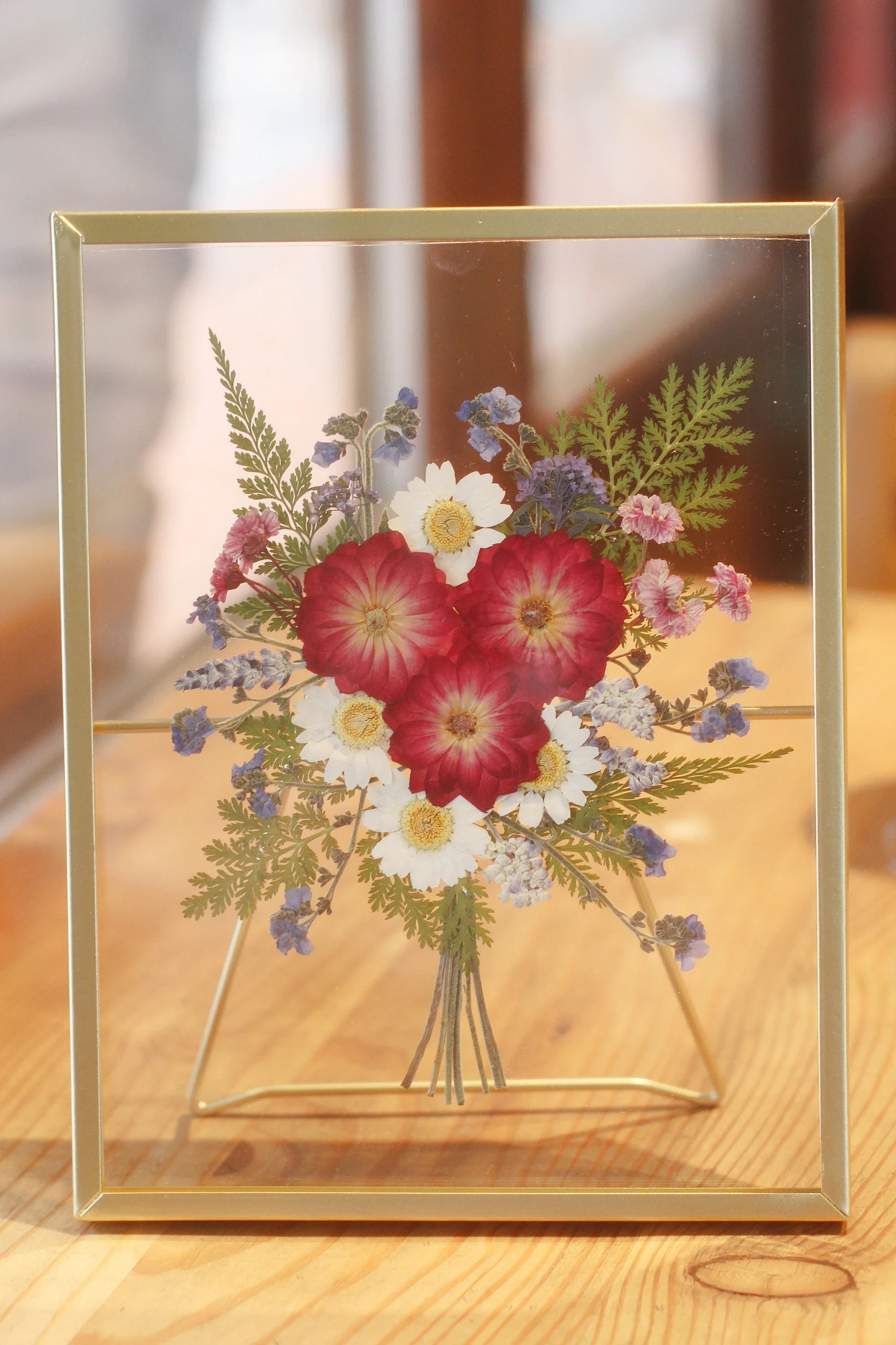 Framed glass display case with a bouquet of mixed flowers inside, including red, white, purple, and pink blossoms, placed on a wooden surface.