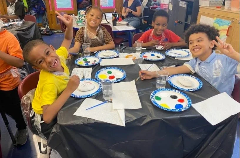 Children celebrating a painting party, sitting at a table with paper, brushes, and paint palettes, smiling and making peace signs.
