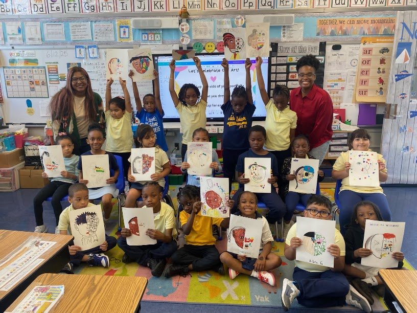 Elementary school classroom with children holding artwork, two teachers standing, colorful educational posters, and a digital screen.