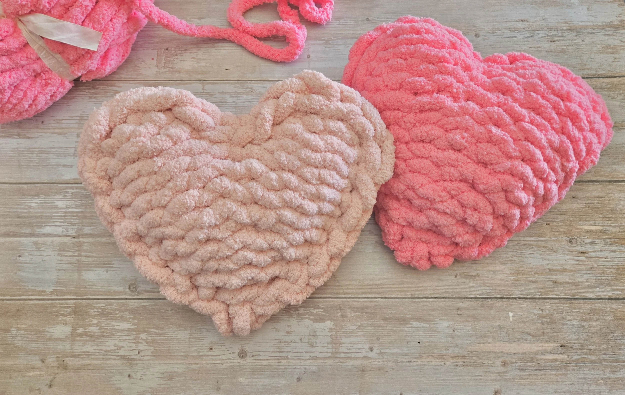 Two plush heart-shaped pillows, one pink and the other light pink, placed on a wooden surface with additional plush items partially visible in the background.