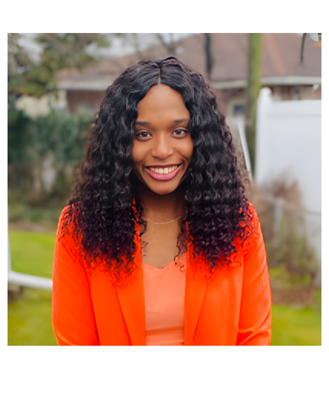 A young woman with curly black hair, smiling, wearing an orange blazer and coral top, standing outdoors with a blurred background of trees and a house.
