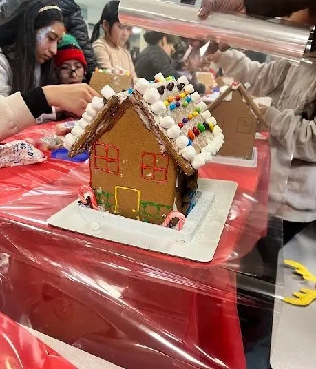 A gingerbread house decorated with candies and icing on a white tray, with children and adults in the background at a holiday event.
