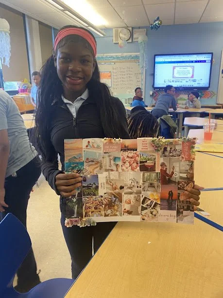 A young girl with long dark hair and a pink headband holding a collage of photographs in a classroom.