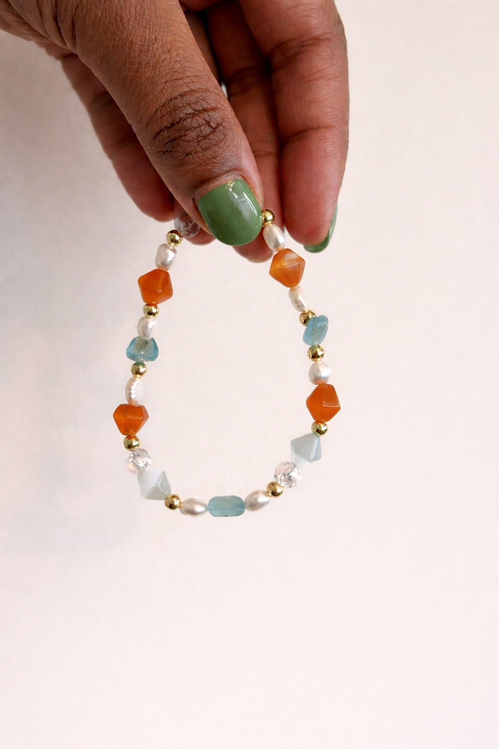 A person's hand with green painted nails holding a colorful beaded bracelet made of various beads, including orange, light blue, white, and gold, against a plain white background.
