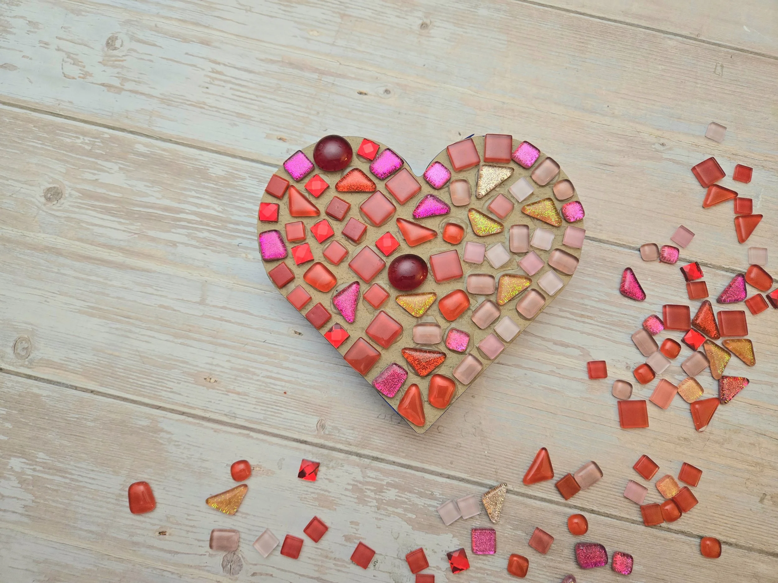 A heart-shaped mosaic made of red, pink, orange, and gold glass tiles, with some scattered pieces on a light wooden surface.