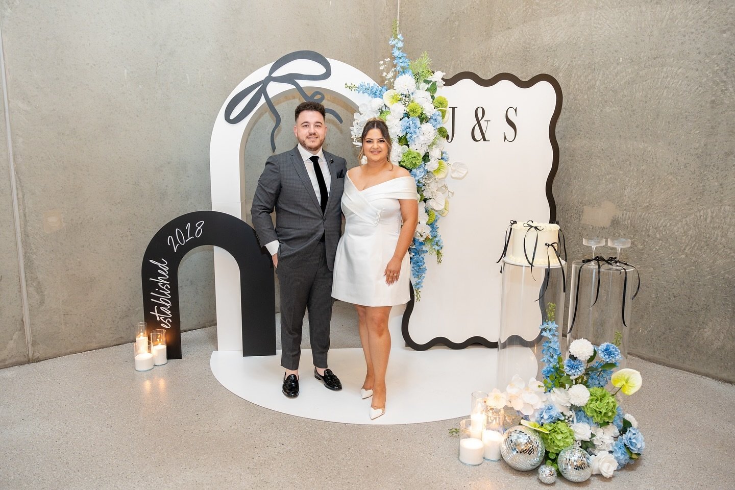 A man and woman standing together at a wedding celebration, in front of a white backdrop with floral arrangements and decorative elements, including candles and mirrored balls.