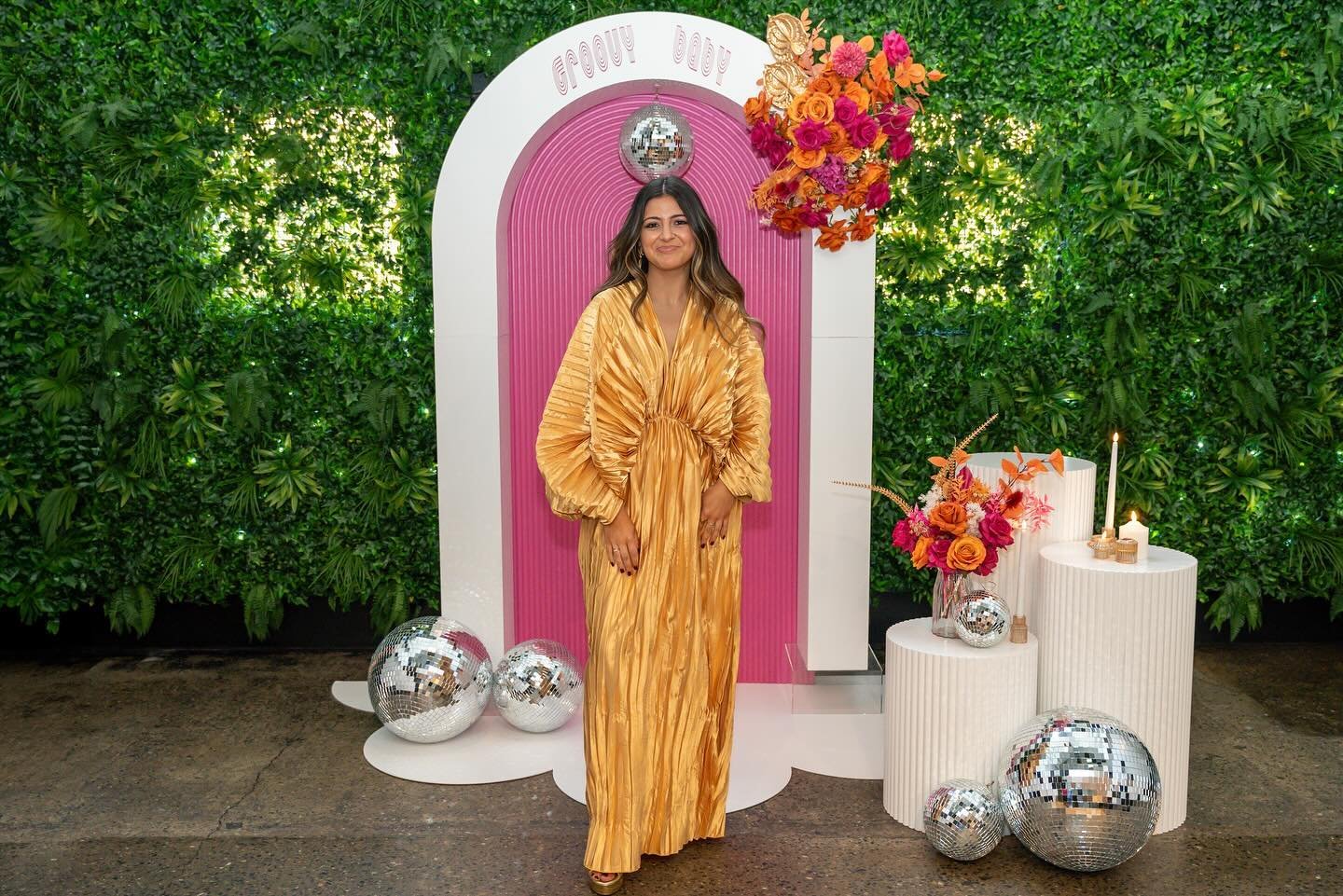 A woman in a long, shiny gold dress stands in front of a pink and white backdrop decorated with colorful flowers and disco balls, with a lush green wall behind her.