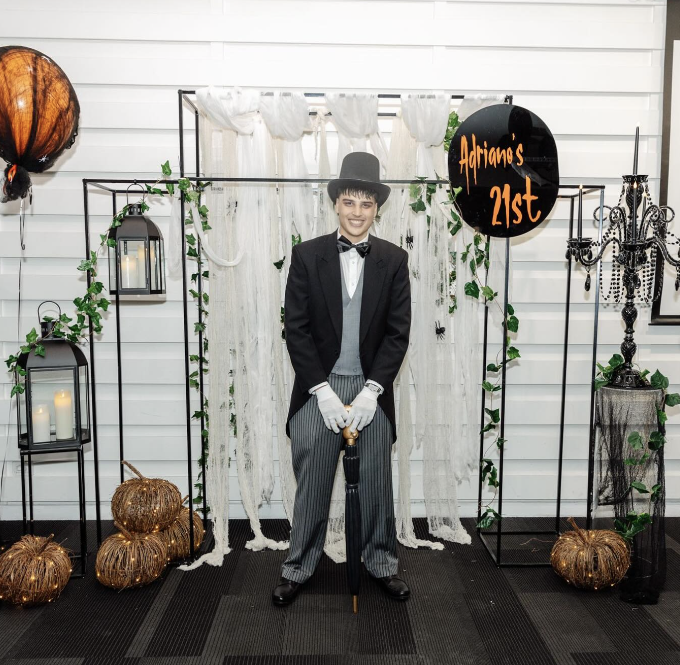 A young man dressed as a vintage magician or ringmaster, wearing a black top hat, black tuxedo jacket, white shirt, gray vest, striped pants, white gloves, and holding a black umbrella, stands in front of a Halloween-themed backdrop with pumpkins, ca