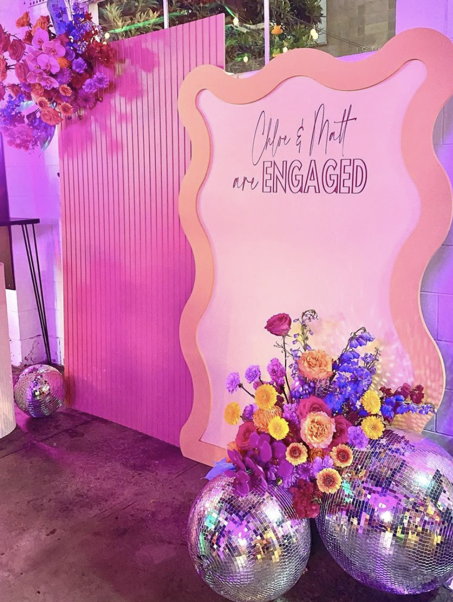 Decorative sign with the words 'Chloe & Matt are ENGAGED,' surrounded by pink and purple flowers in mirrored disco ball vases. The background features a pink striped wall and a floral arrangement in a tall vase.