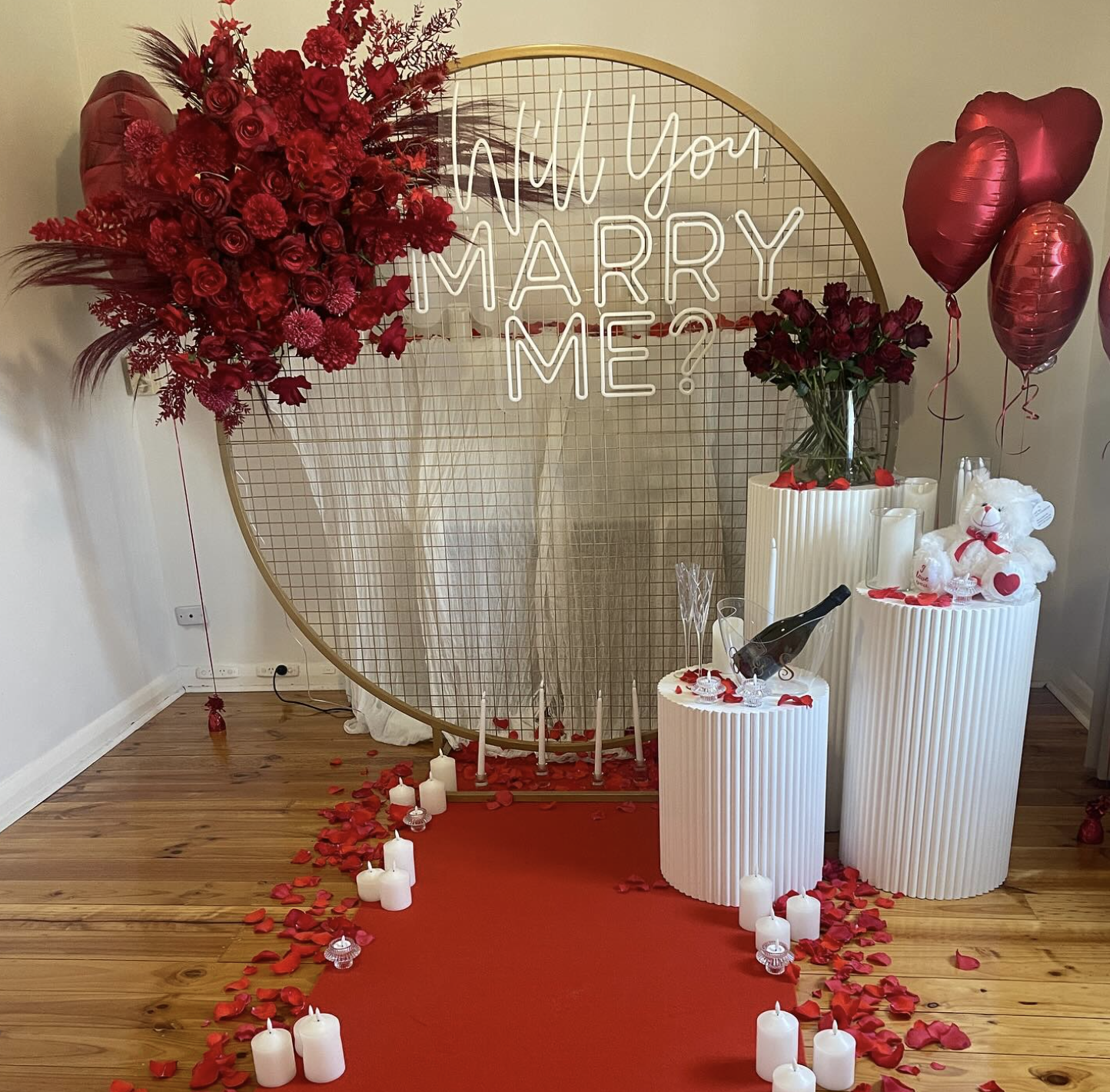 Decorative wedding proposal setup with a large round wire backdrop displaying a neon sign that reads 'Will You Marry Me?' in white, surrounded by red flowers, heart-shaped balloons, candles, roses in a vase, and plush teddy bears.