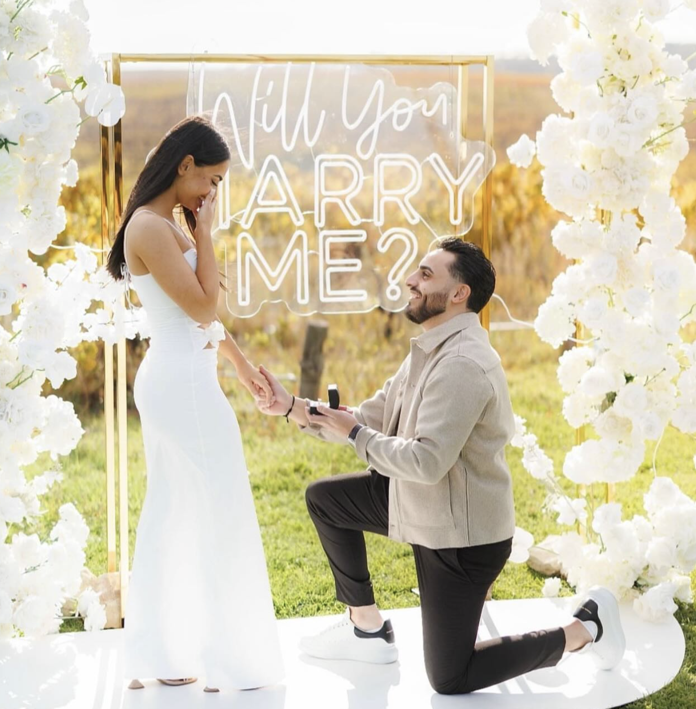 A man proposes marriage to a woman during a daytime outdoor proposal, with a sign in the background that reads 'Will you marry me?' and decorated with white flowers.