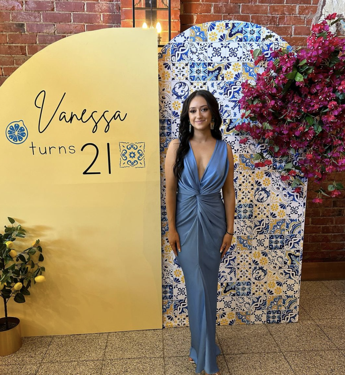 A young woman in a blue dress standing next to a yellow sign that says "Vanessa turns 21" and a decorative blue and yellow tile backdrop with a pink flowering plant nearby.