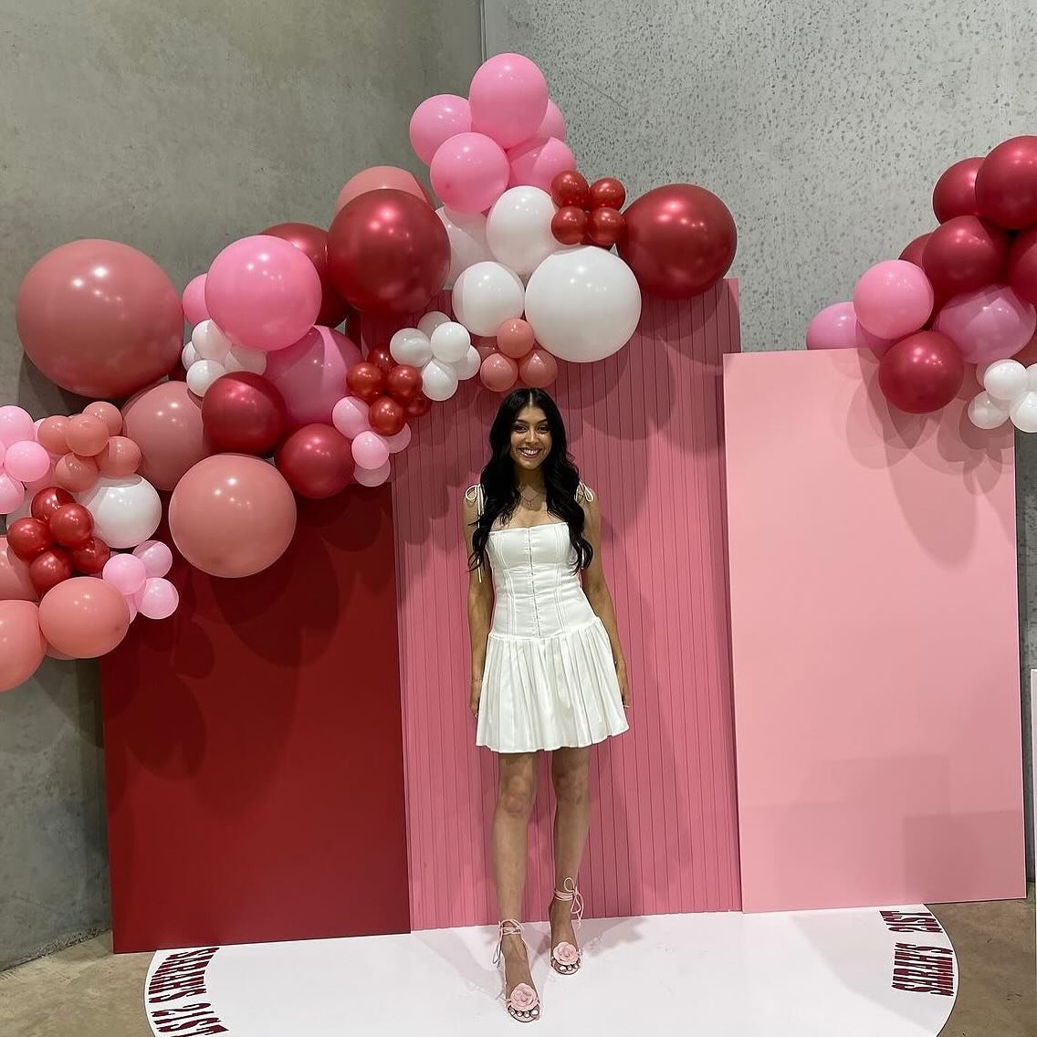 A woman in a white dress standing in front of pink and red balloon decorations and pink wall panels.