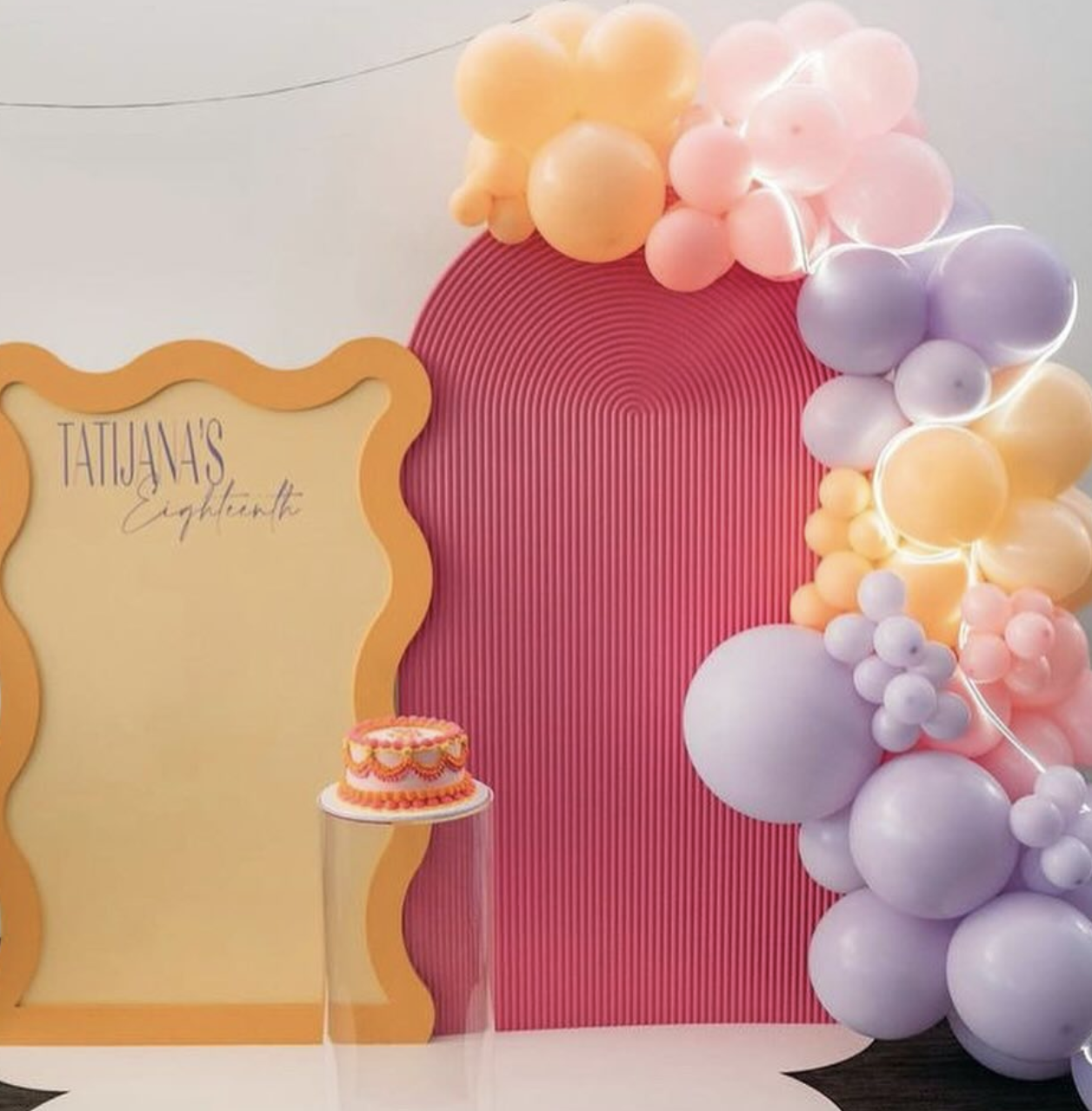 Decorative backdrop with pink, purple, peach balloons arranged on the right side, a pink radially ridged arch, a yellow frame on the left with text, and a small cake on a glass stand in front of the frame.