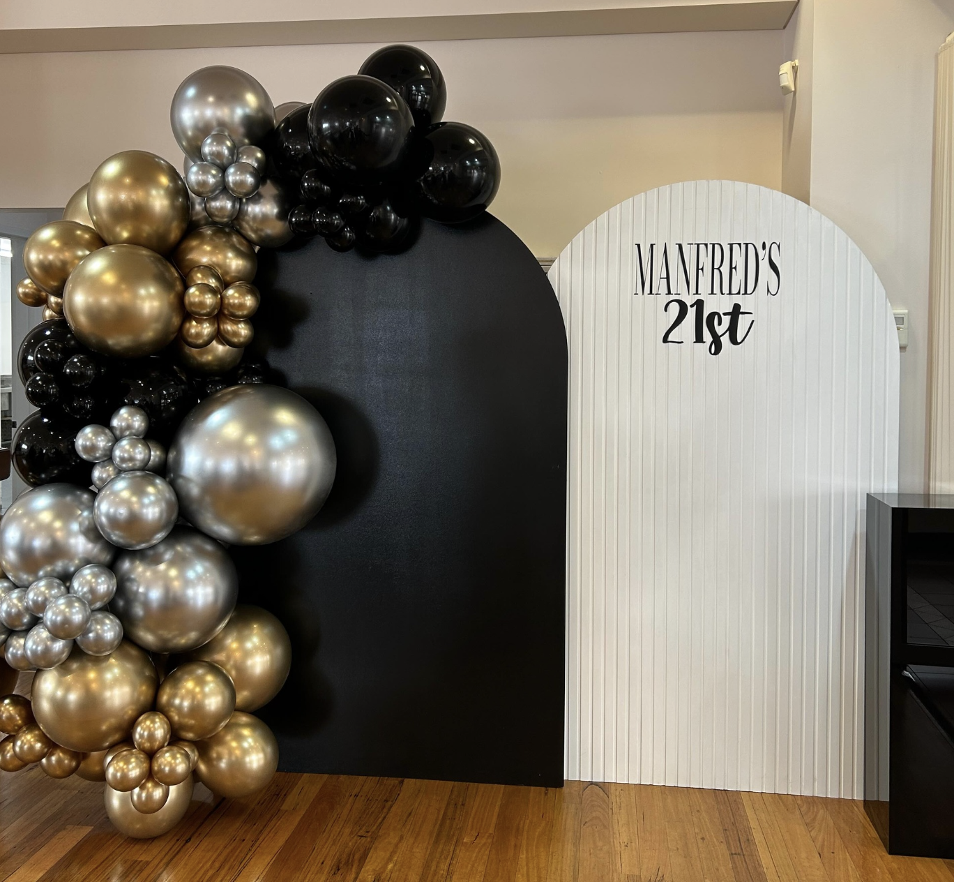 Decorative birthday backdrop with black, gold, and silver balloons on a black and white panel, with the white panel reading 'MANFRED'S 21st'.