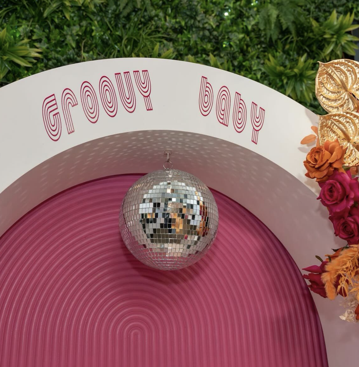 A sign that reads 'froufrou baby' with a pink circular backdrop, a disco ball hanging in the center, and colorful flowers on the right side, with greenery in the background.