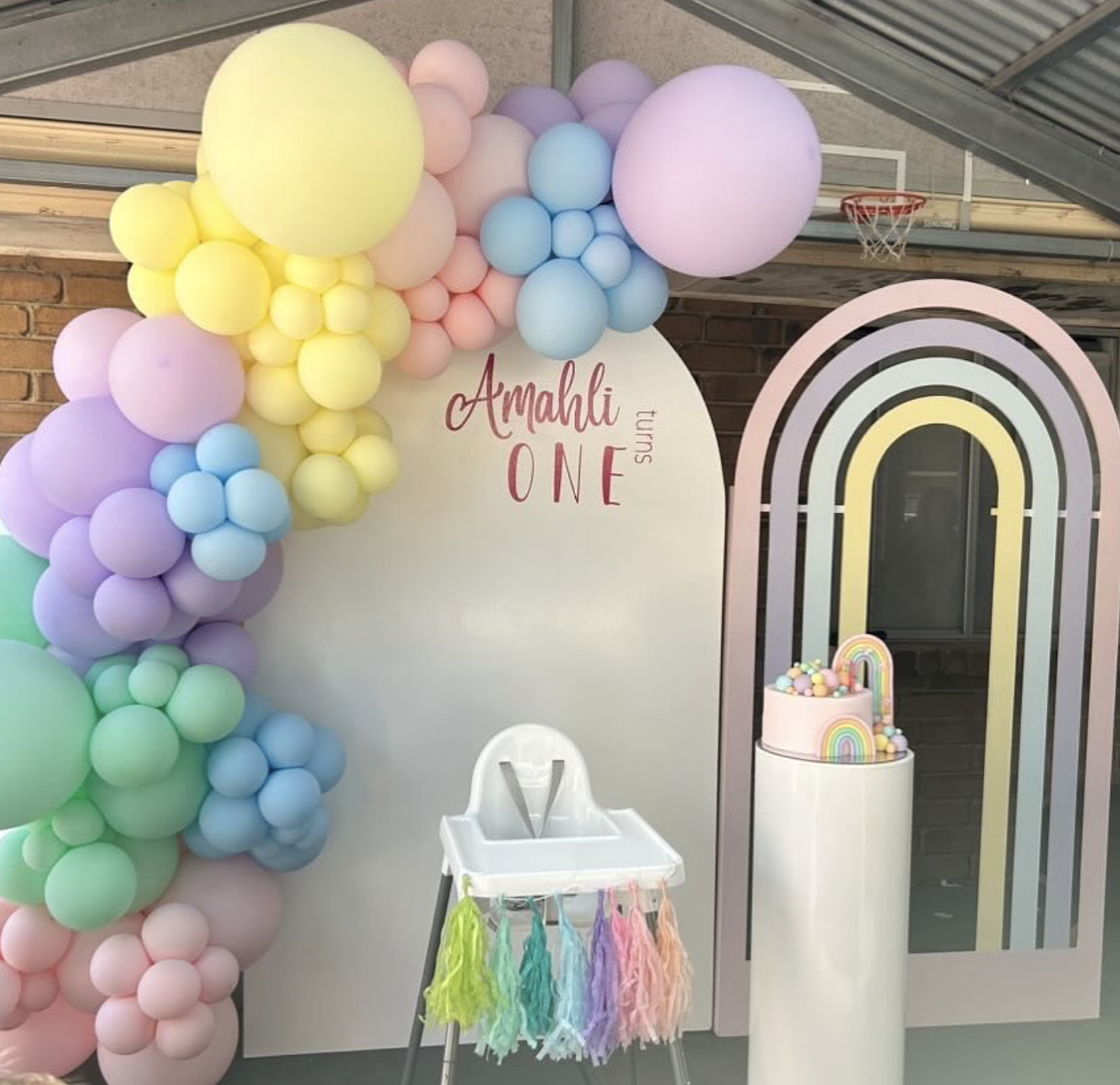 Colorful pastel birthday decorations with balloon garland, a white backdrop with the words 'Amarli Turns One', a rainbow-themed cake, and a high chair with rainbow tassels.