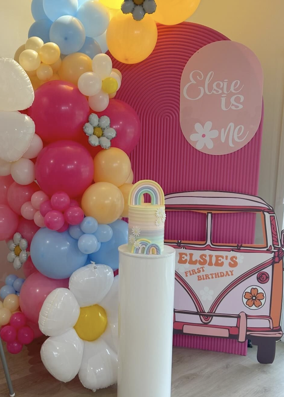 Decorative backdrop for Elsie's first birthday featuring a balloon arrangement with pink, blue, white, and yellow balloons, a pink background with a floral sign that says 'Elsie is me', and a cutout of a pink vintage van with 'Elsie's First Birthday'