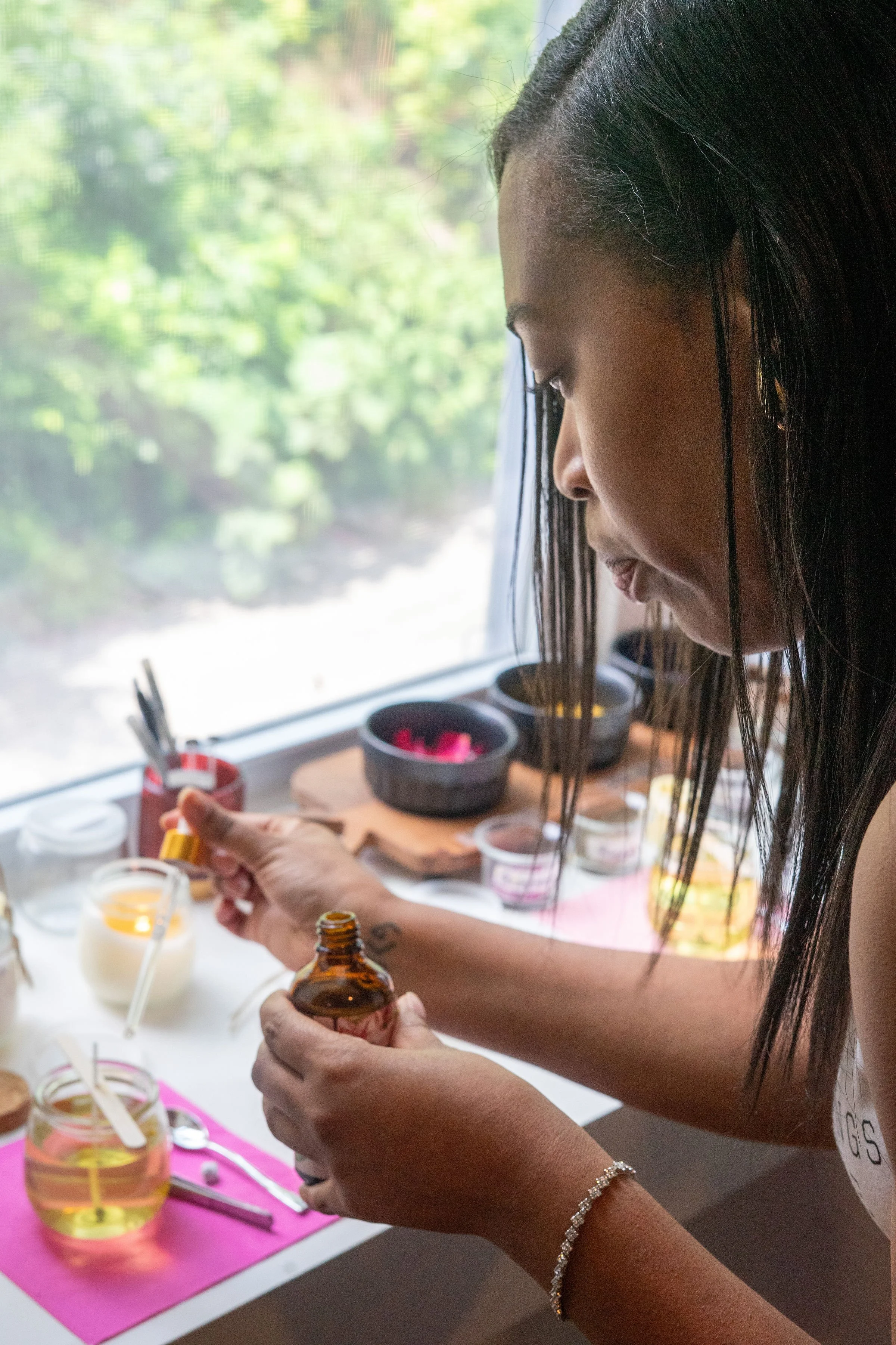 A woman with dark hair applying essential oil or perfume from a small amber bottle, surrounded by jars on a white counter with a pink mat. The background includes a window with greenery outside.