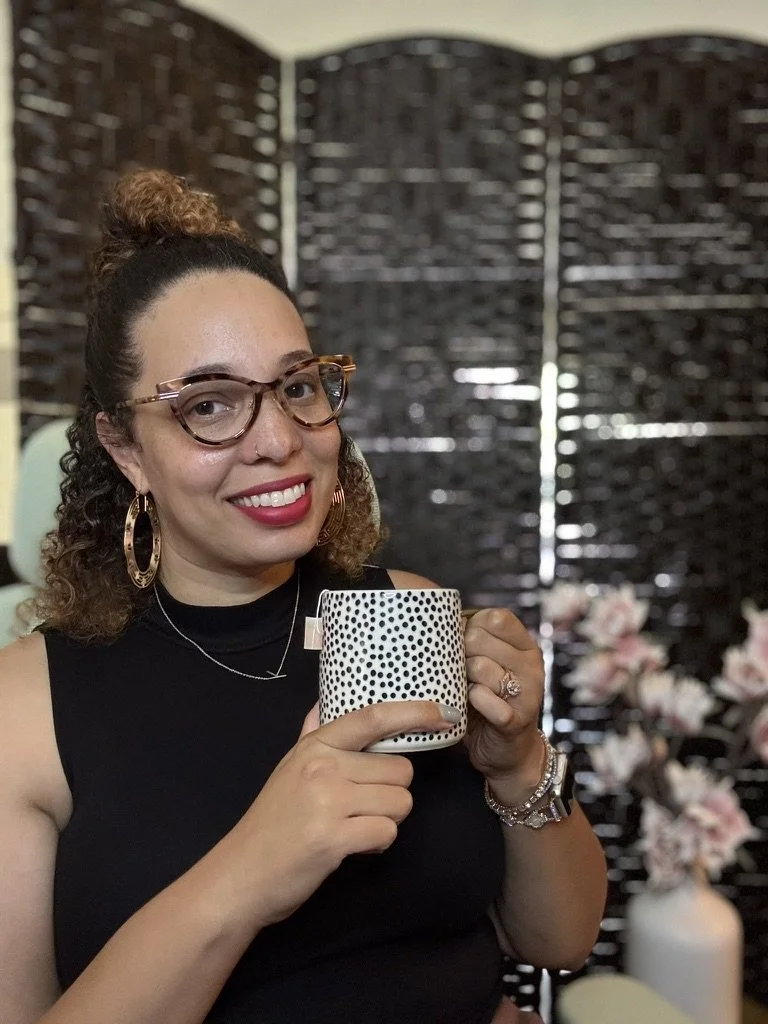 Natashja, a woman smiling at her desk with glasses and red lipstick and a black top holding a polkadot mug with tea and flowers in the background