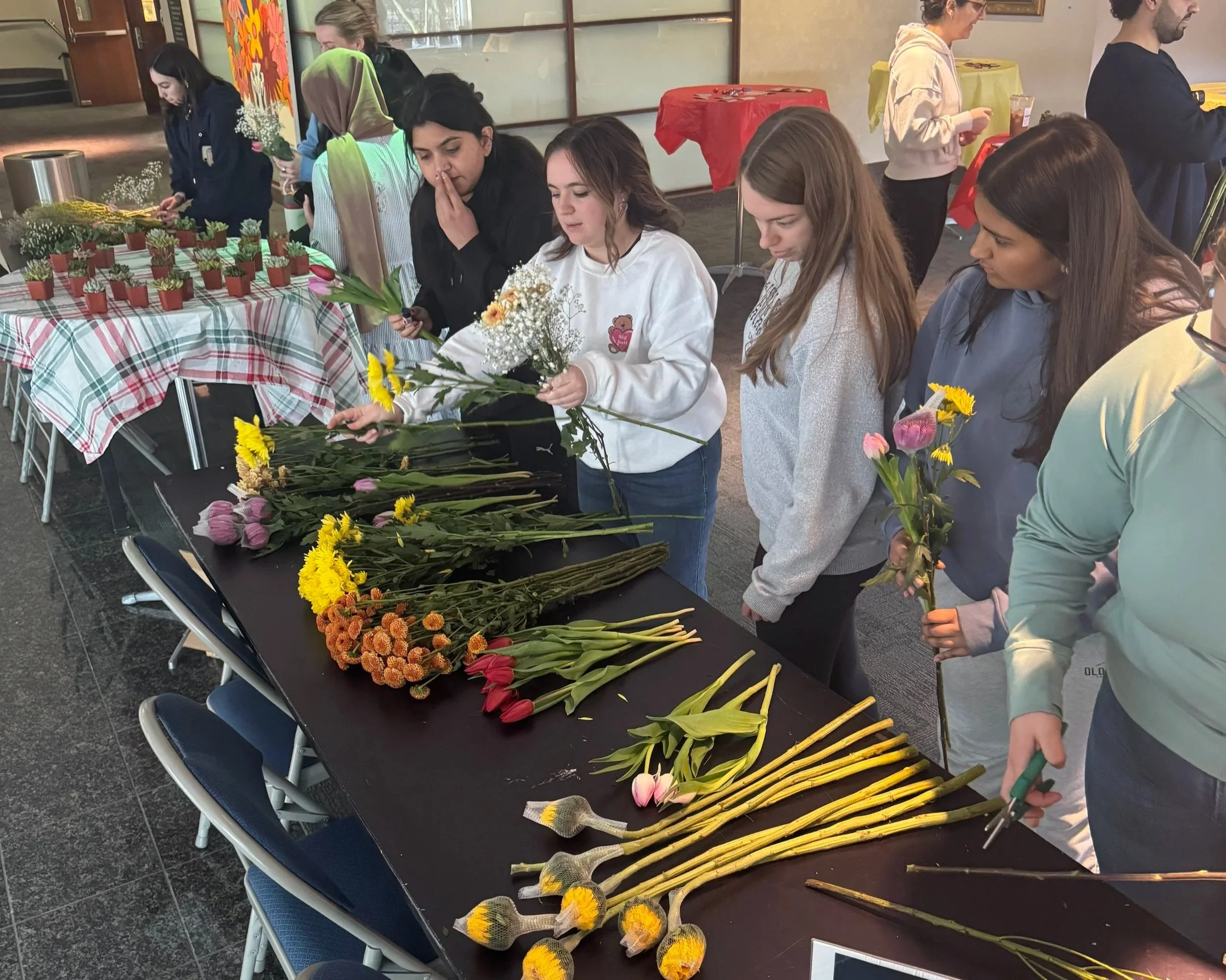 People gathered around a table with flowers, arranging and holding various types of fresh flowers including tulips, sunflowers, and baby's breath.
