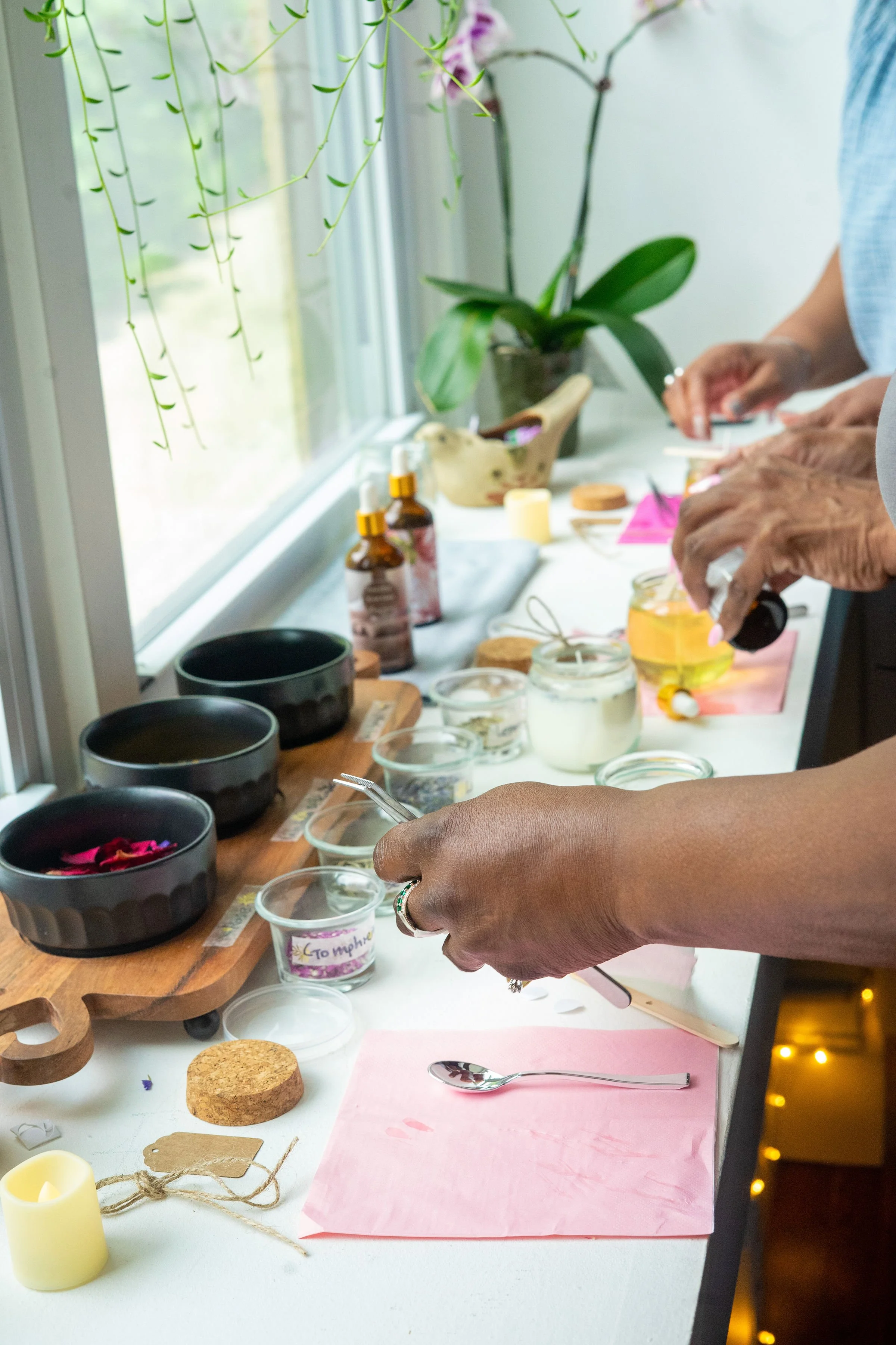 People arranging filling small jars with wax and oils on a white table near a window with potted orchids and skincare bottles. Sacred Scents and Sounds Wellness Event