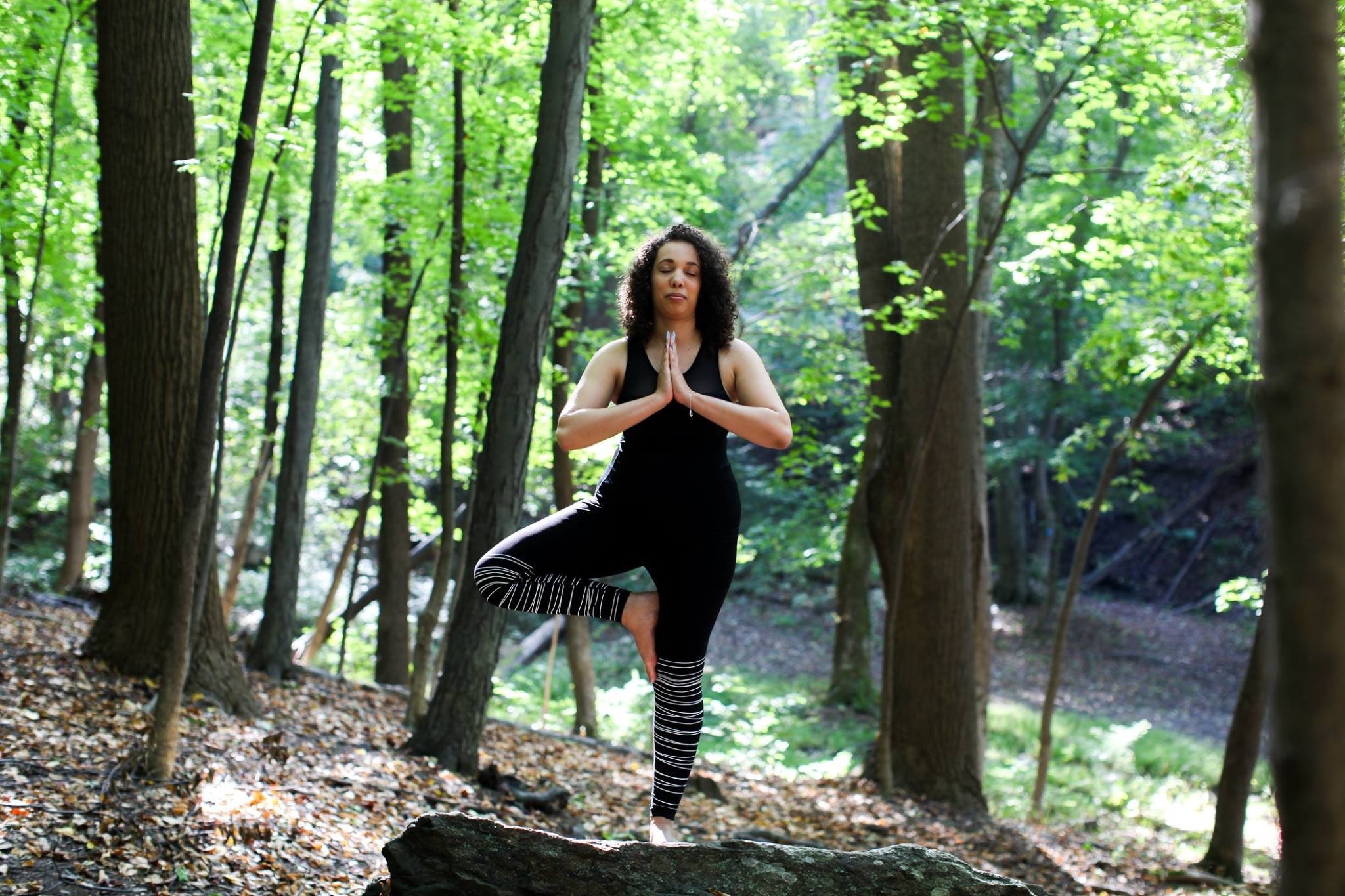 A woman practicing yoga in a forest, standing on one leg in a tree pose with hands together in prayer position.