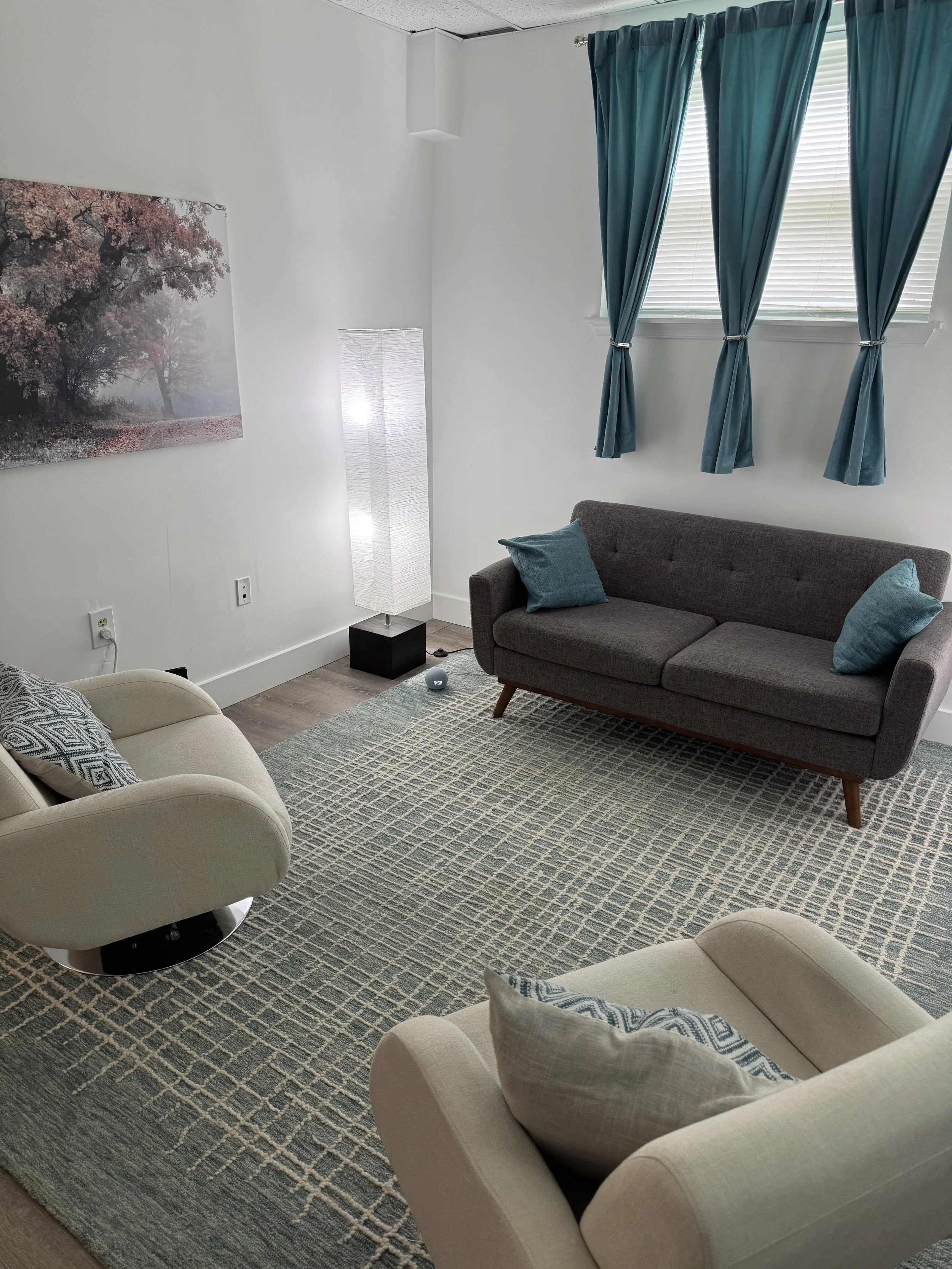 A cozy living room with a beige armchair and a gray sofa, both adorned with blue patterned pillows. A patterned rug covers the wooden floor, and a tall white floor lamp and a painting of trees with pink leaves decorate the white wall. A window with multiple blue curtains and blinds lets in natural light.