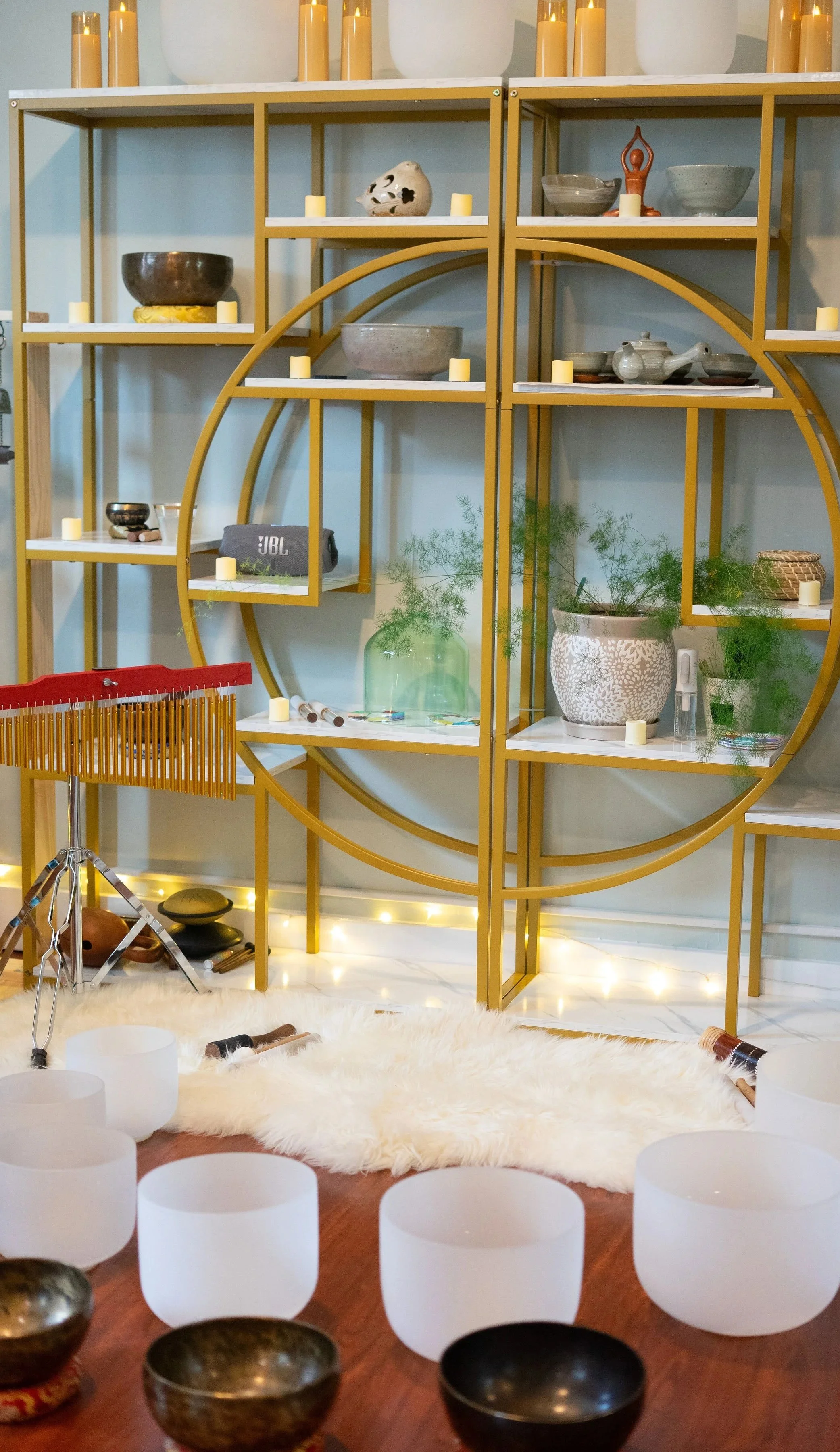 A decorative shelving unit with various bowls, candles, and plants, illuminated by string lights at the base, with singing bowls and meditation chimes in the foreground on a wooden floor, and surrounded by white candles and a furry rug.