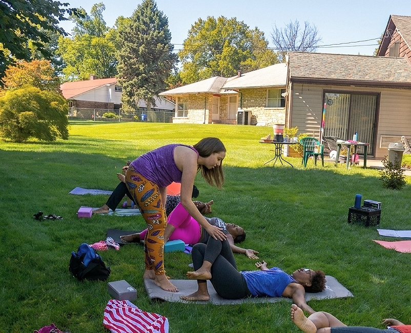 A woman practicing yoga outdoors on a grassy lawn with adults, in a suburban backyard setting with houses, trees, and outdoor furniture in the background.