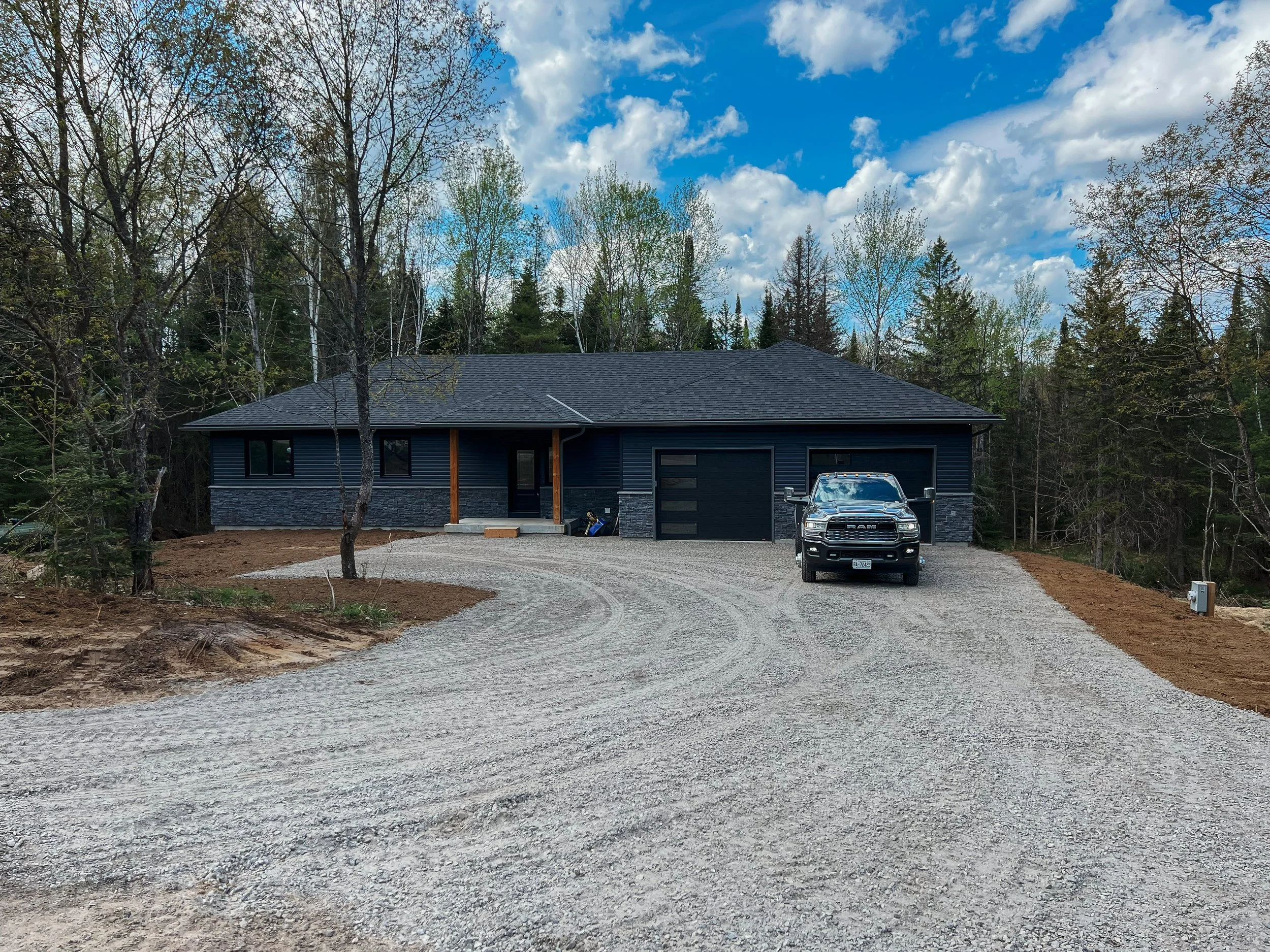 Newly built house with dark exterior, stone accents, a driveway, and a black pickup truck parked in front, surrounded by trees and a partly cloudy sky.
