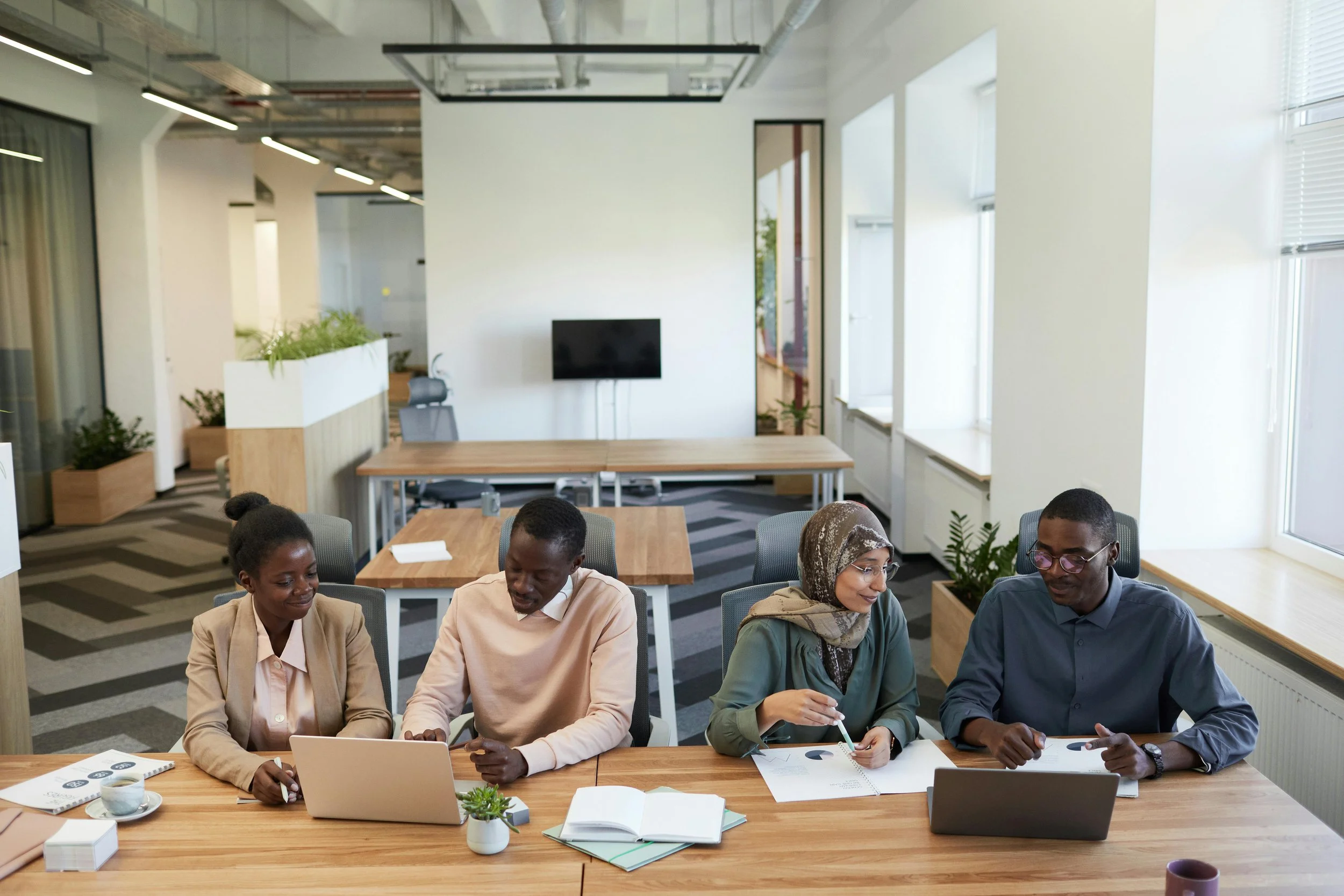 A diverse group of people, including women and men, sitting at a long conference table in a modern office, engaged in work and discussion. The office has large windows, plants, a TV on the wall, and natural lighting.