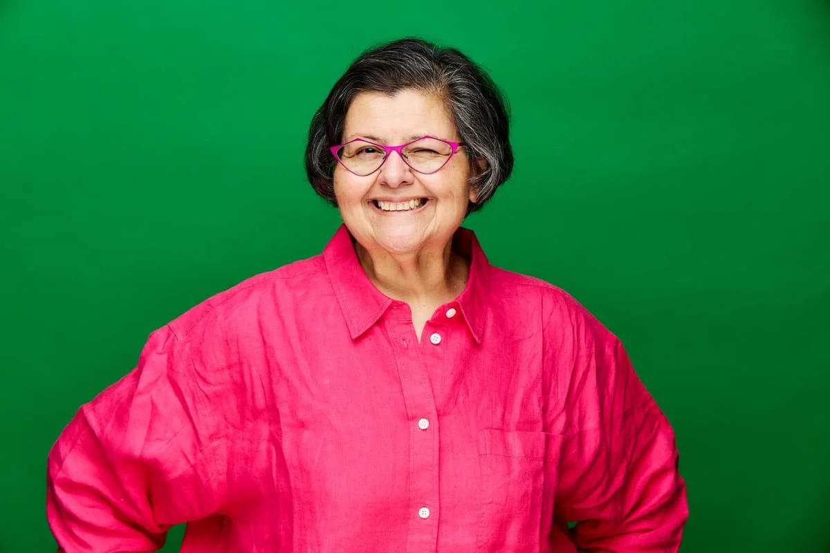 a smiling woman with black hair wearing glasses, a bright pink collared shirt, with her hands on her hips
