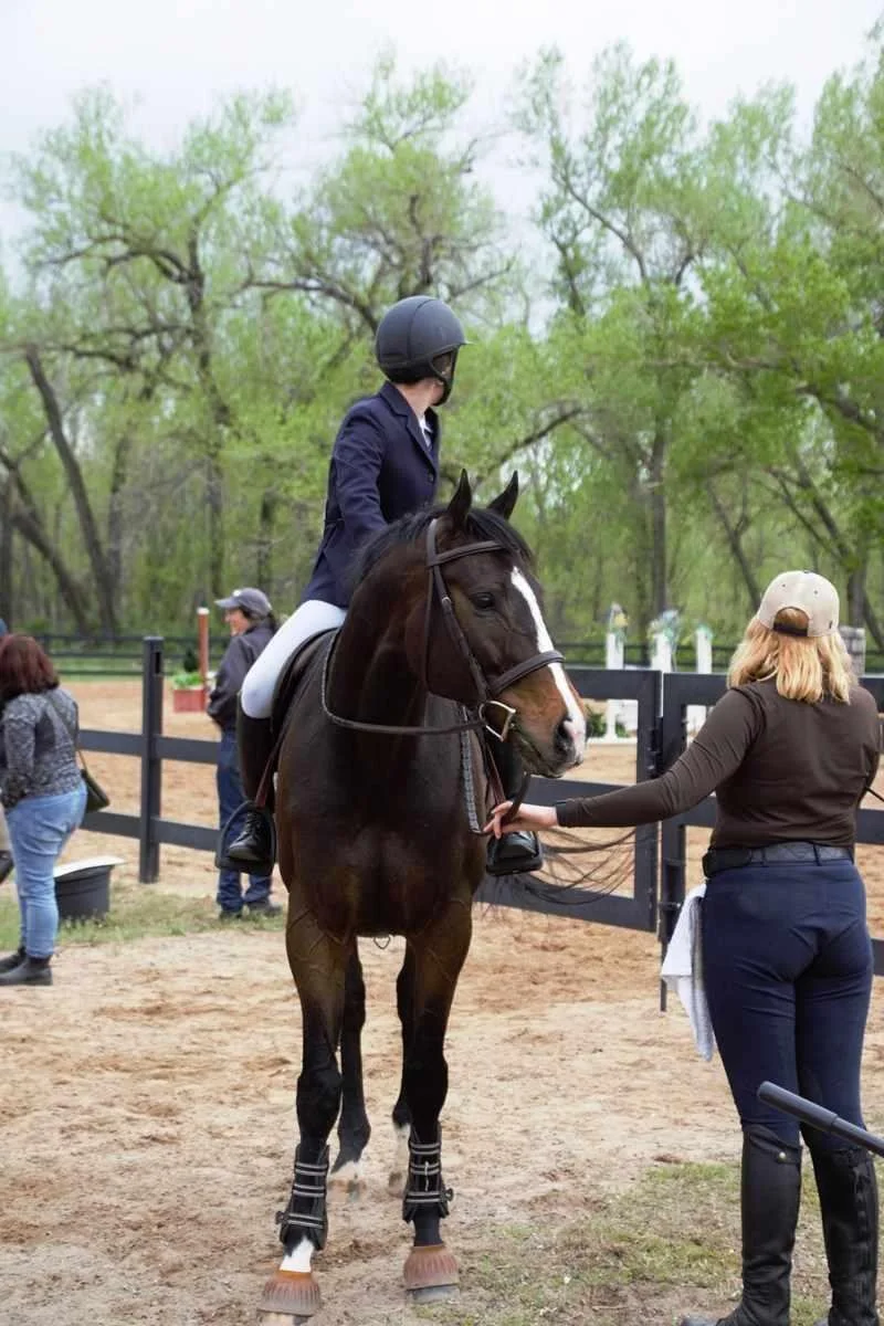 A person in riding attire and a helmet is sitting on a brown horse with white markings on its face. A woman standing beside the horse is holding the reins.