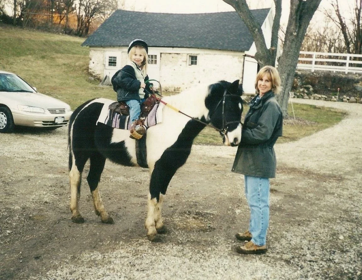 A young girl sitting on a black and white pony with an adult woman standing next to the pony, holding the reins outdoors in a rural setting with trees, a small building, a car, and a grassy hillside in the background.