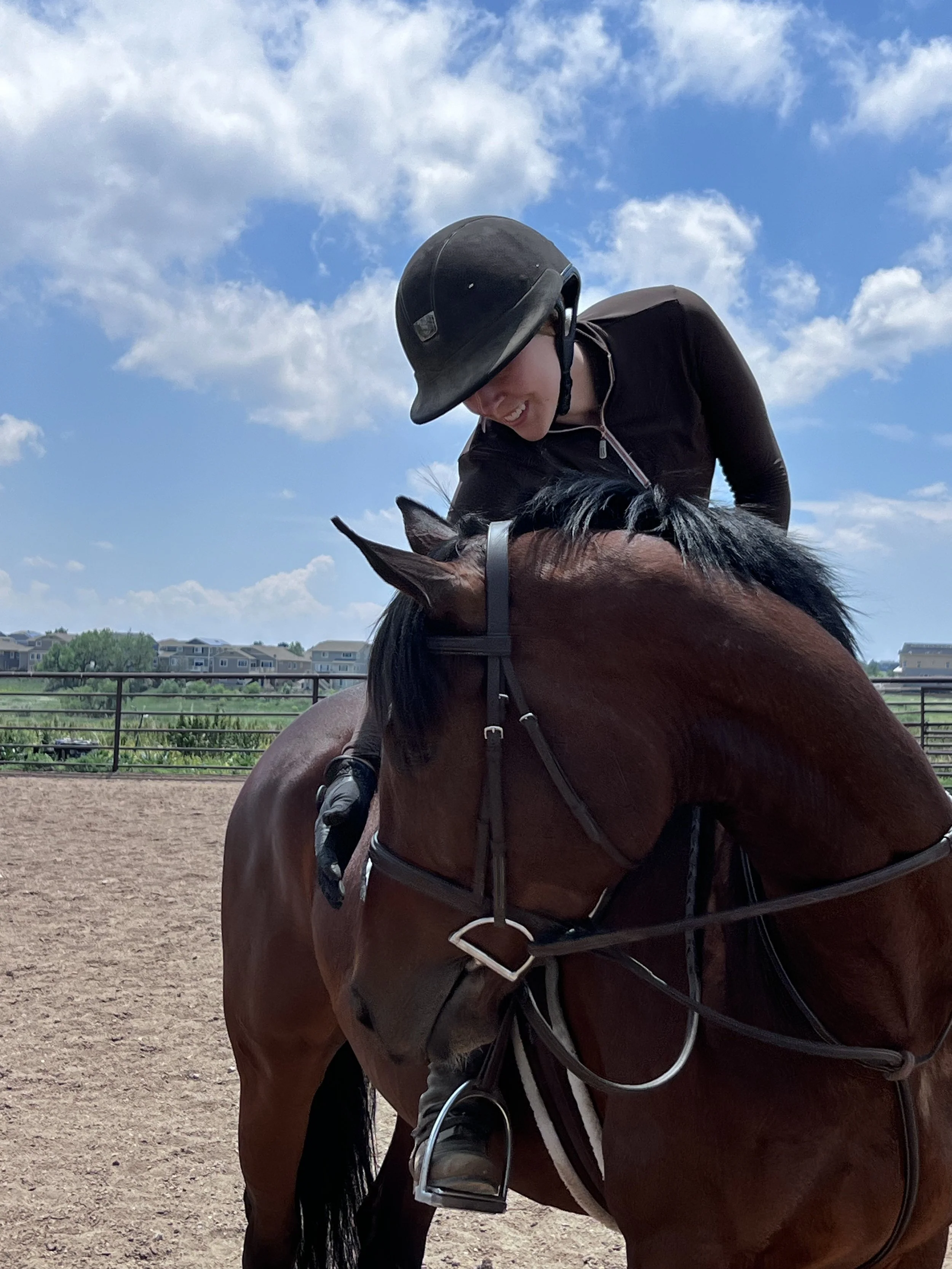 A person wearing a black helmet and black riding jacket leaning over a brown horse with black mane in an outdoor riding arena, smiling. There are houses and green trees in the background under a partly cloudy sky.