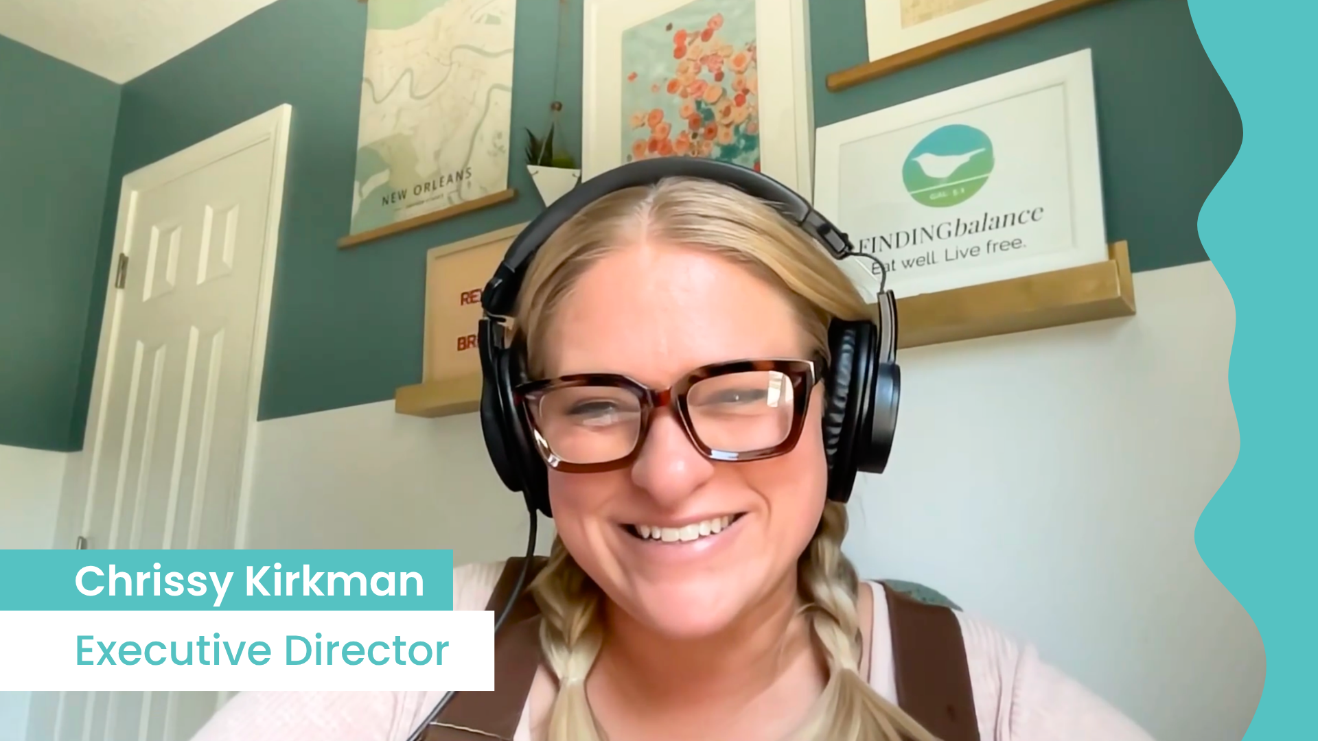 A smiling woman with blonde hair in braids wearing glasses and large headphones, sitting in a room with green and white walls decorated with artwork and a shelf with a framed logo that says "FINDING Balance, Eat well. Live free."