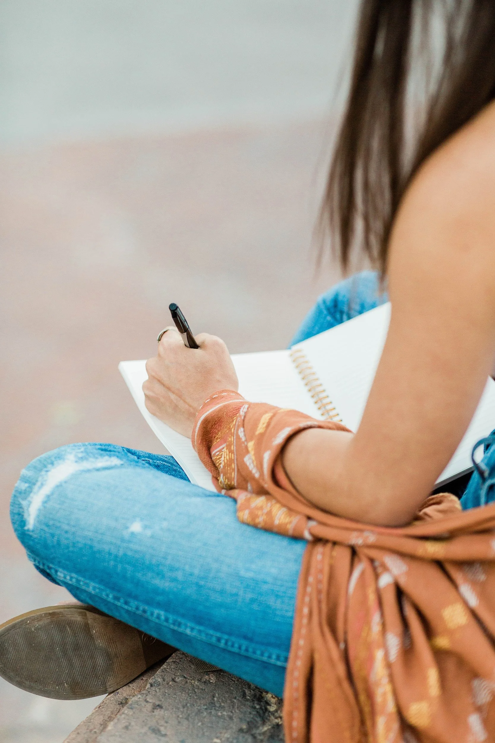 Person sitting cross-legged on a bench, writing in a spiral notebook with a black pen, wearing a brown patterned shirt and blue jeans.