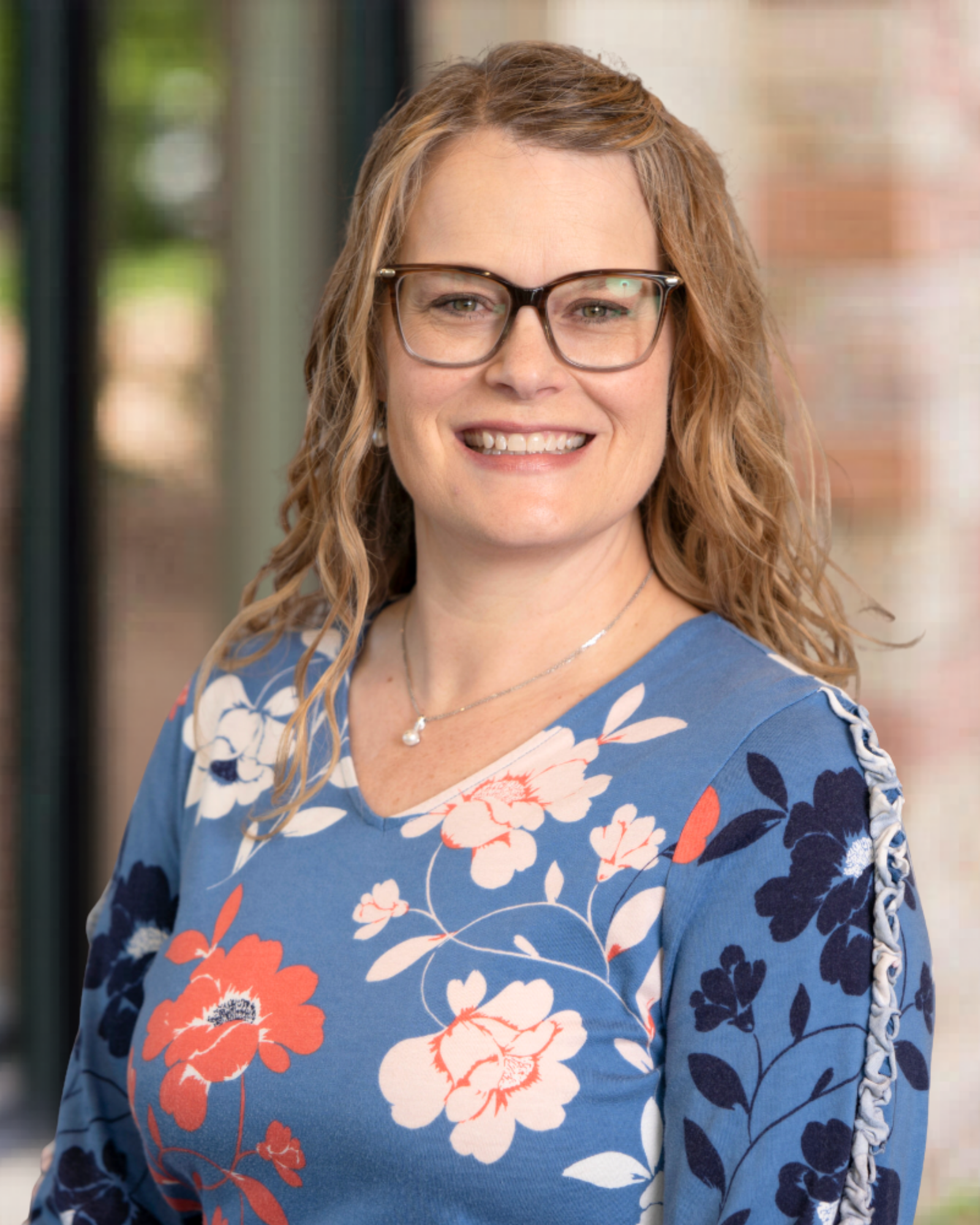 A woman with light brown hair and glasses smiling outdoors, wearing a navy blouse with pink floral patterns and a silver necklace.