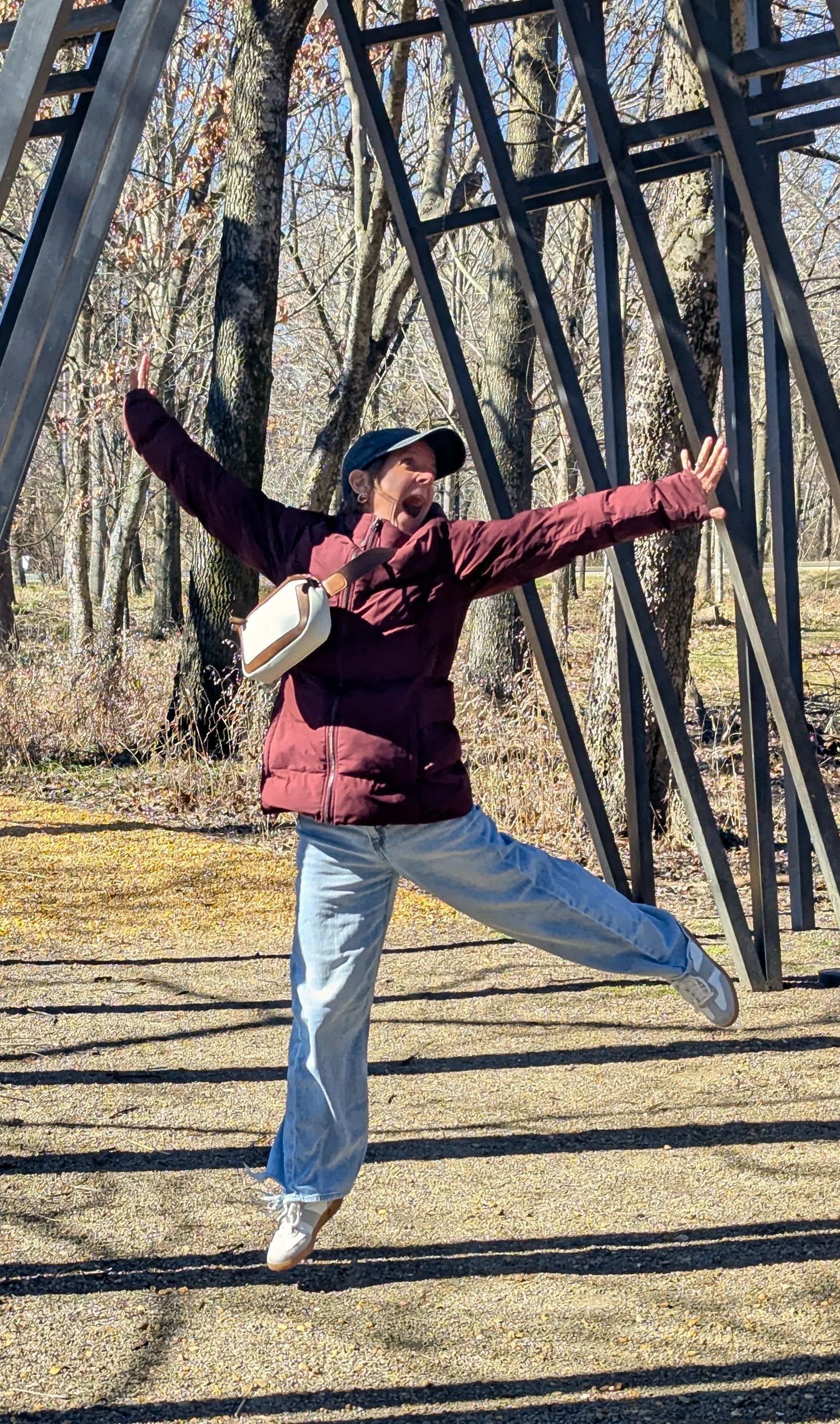 Person in a burgundy jacket, light blue pants, and a cap striking a joyful pose outdoors among trees and shadows, with metal sculptures and bare trees in the background.