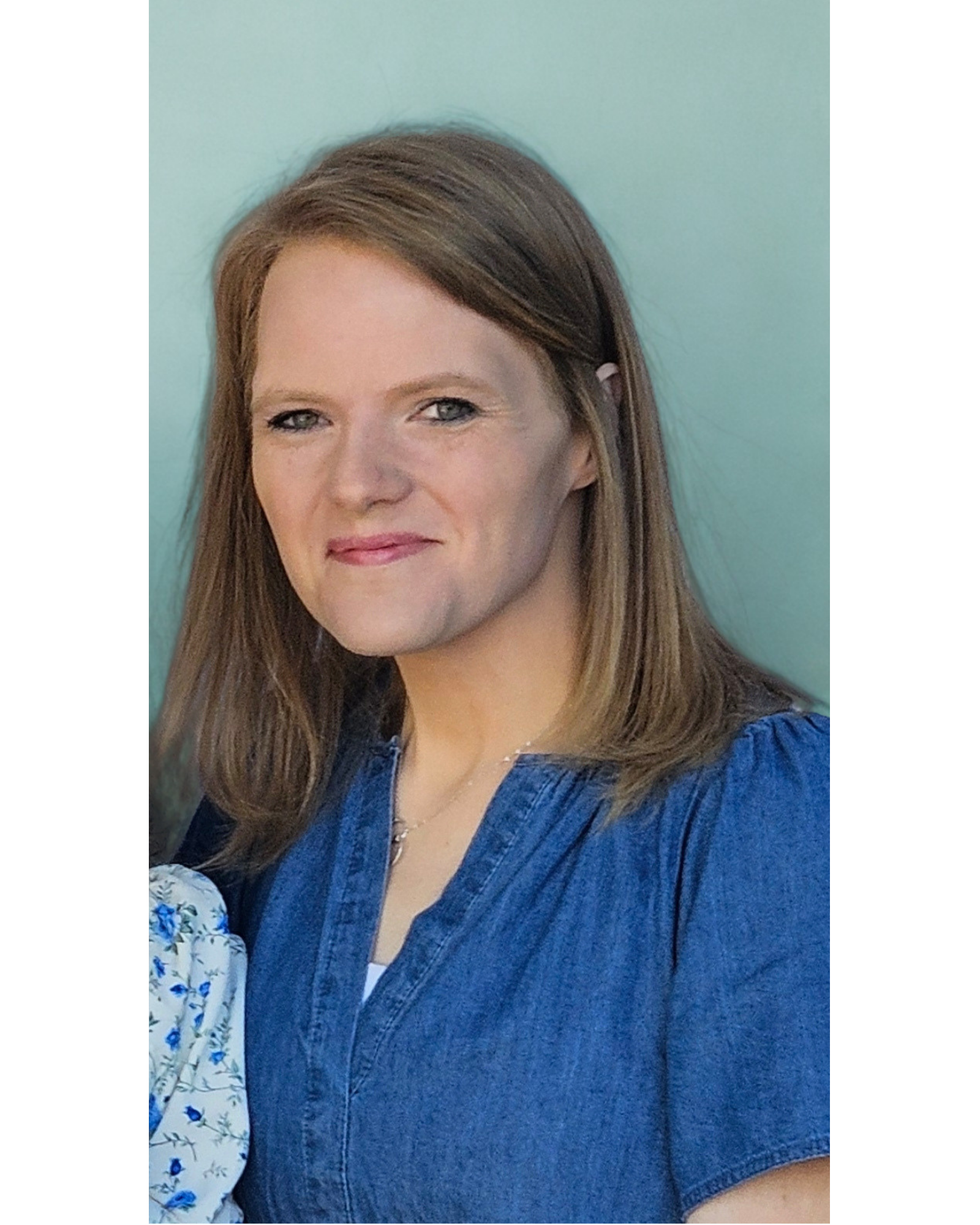 A woman with brown hair and blue eyes smiling, wearing a blue dress with a white floral pattern on the sleeves, standing against a light turquoise background.