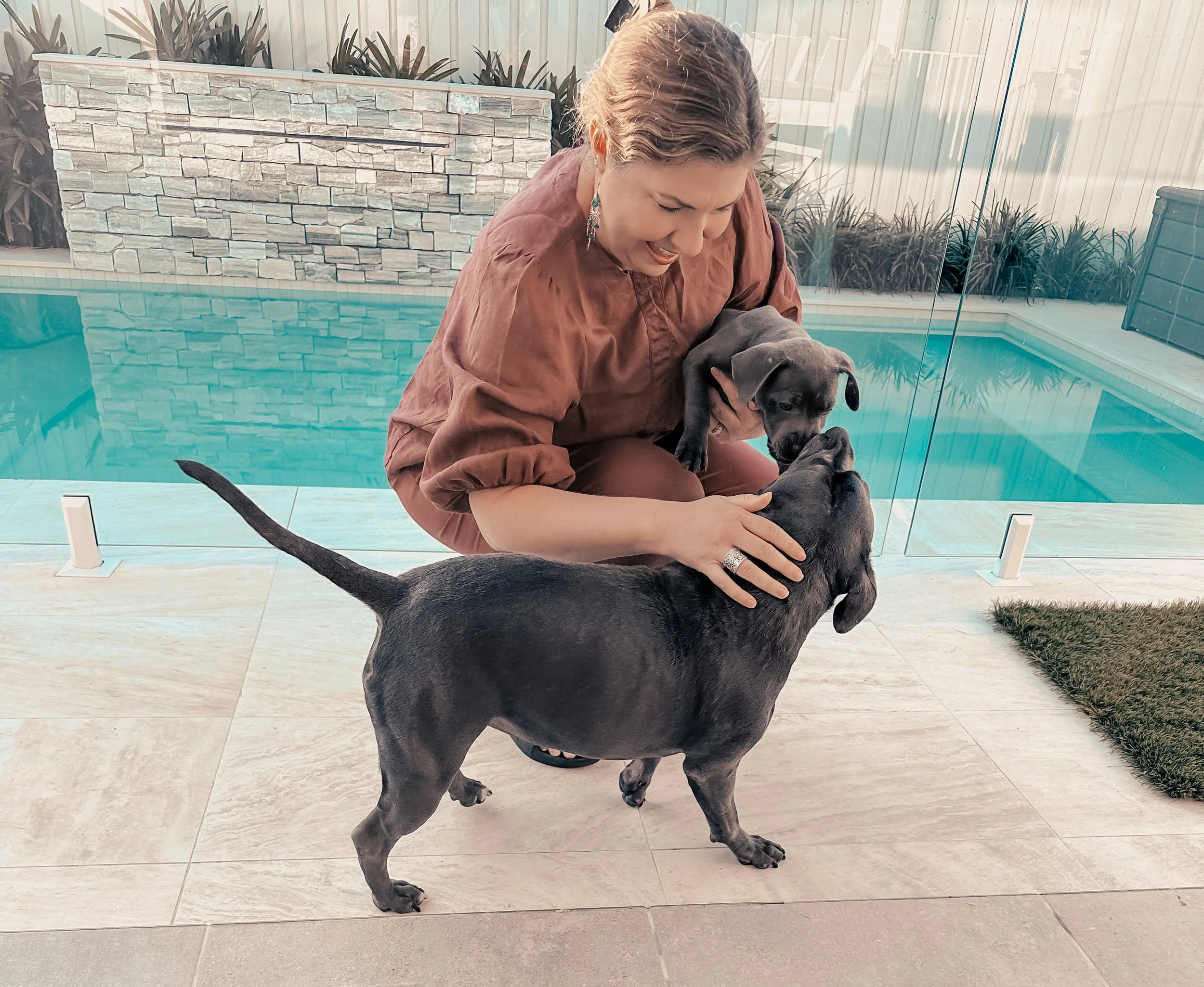 A woman with earrings holding a black puppy and a larger black puppy standing on the floor in front of her, inside a space with a swimming pool, glass wall, and plants in the background.