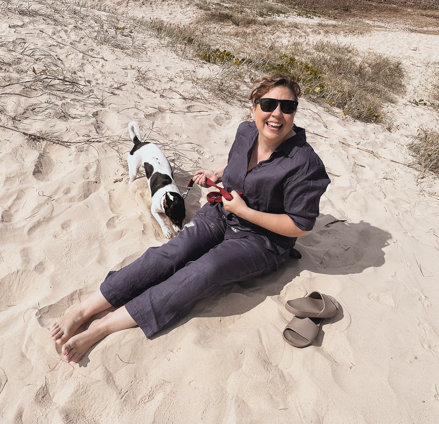 A woman with short, wavy hair, sunglasses, and a dark outfit sitting on sandy beach with a small black and white dog on a leash. There are some plants and a shadow on the sand, with a pair of gray slip-on shoes nearby.